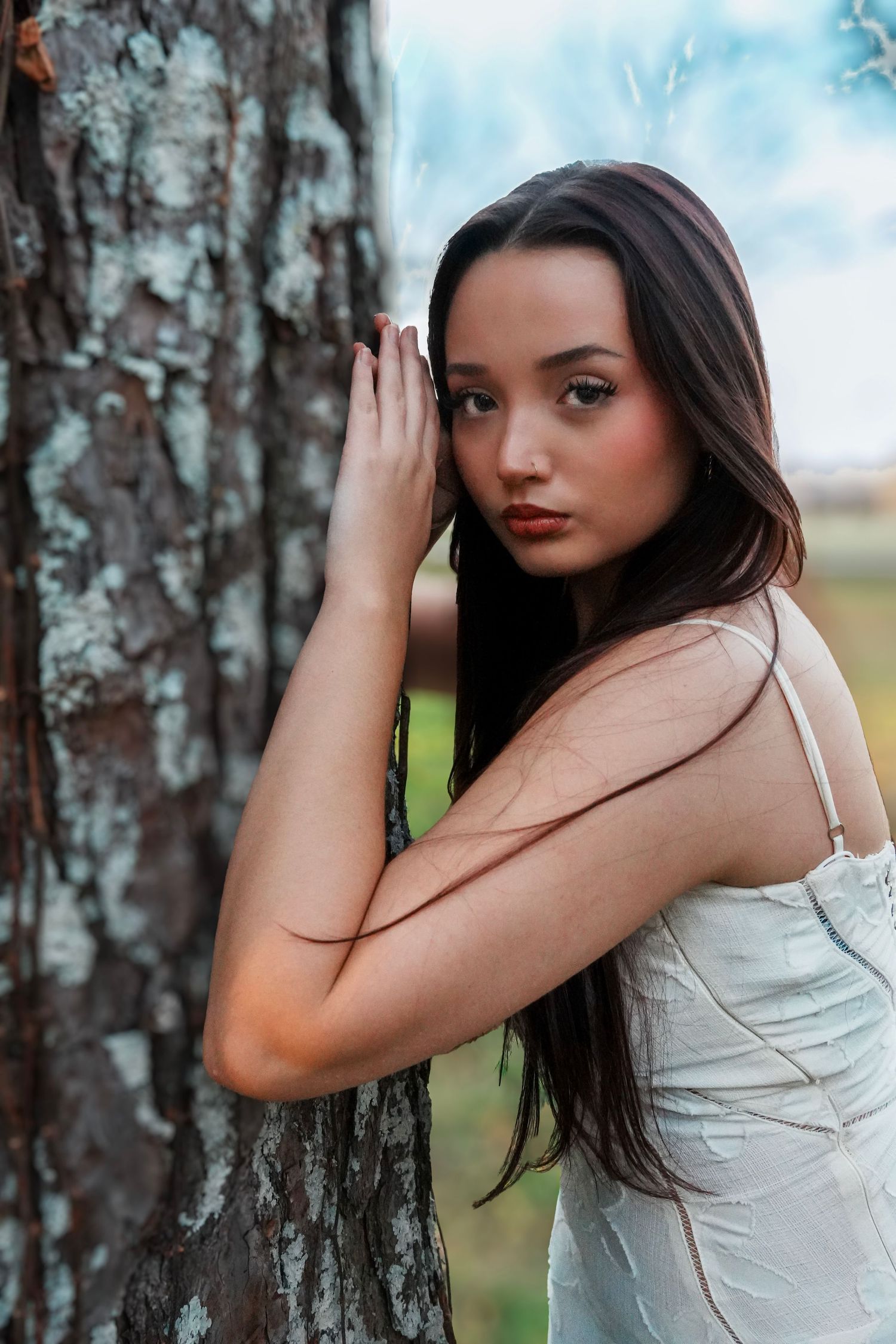 Brunette woman in white dress leans against tree bark in serene outdoor portrait.