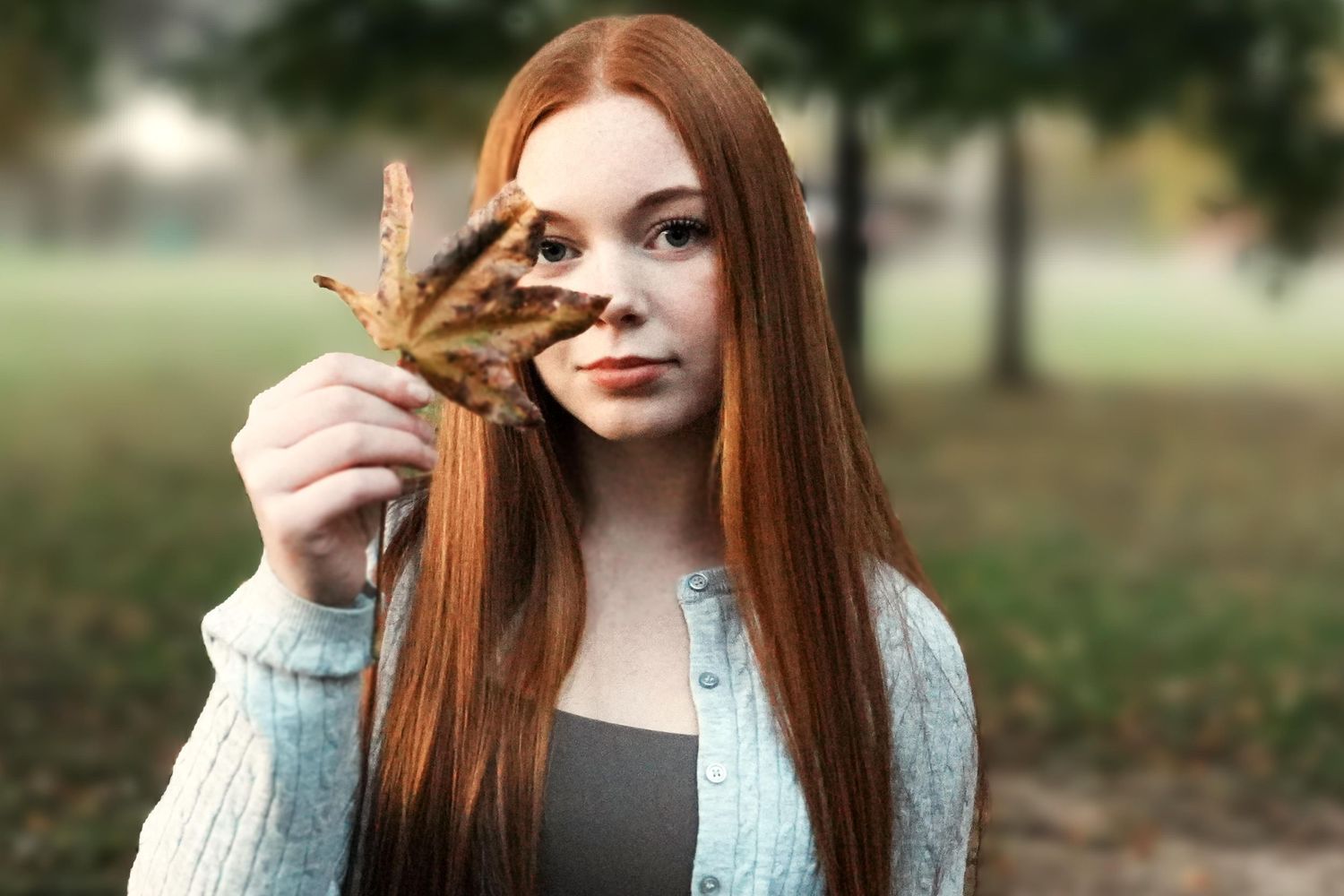 A young woman with long red hair holds an autumn leaf outdoors in a park setting.