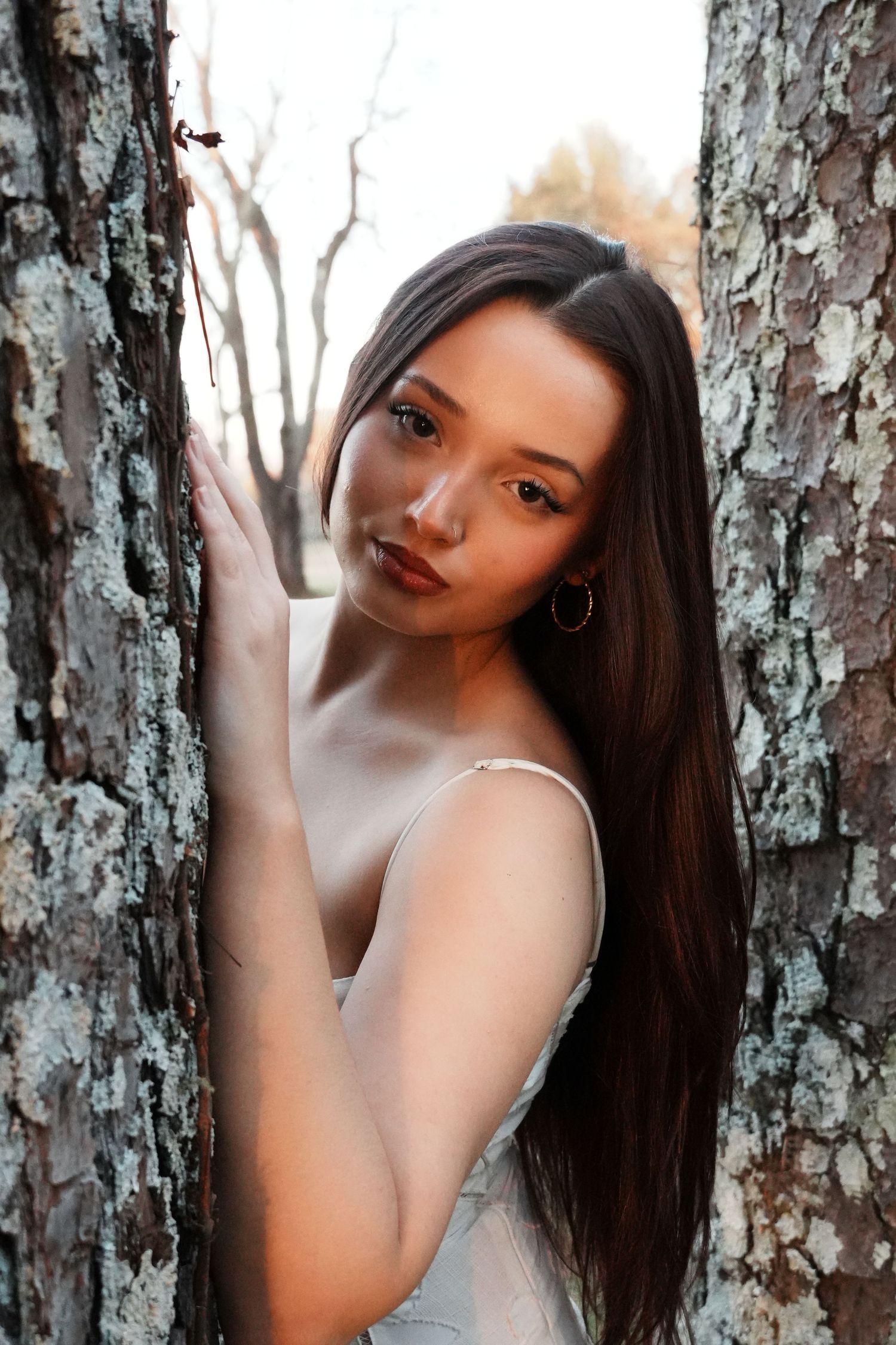 Young brunette woman in silver slip dress posing between two lichen-covered tree trunks in autumn park.