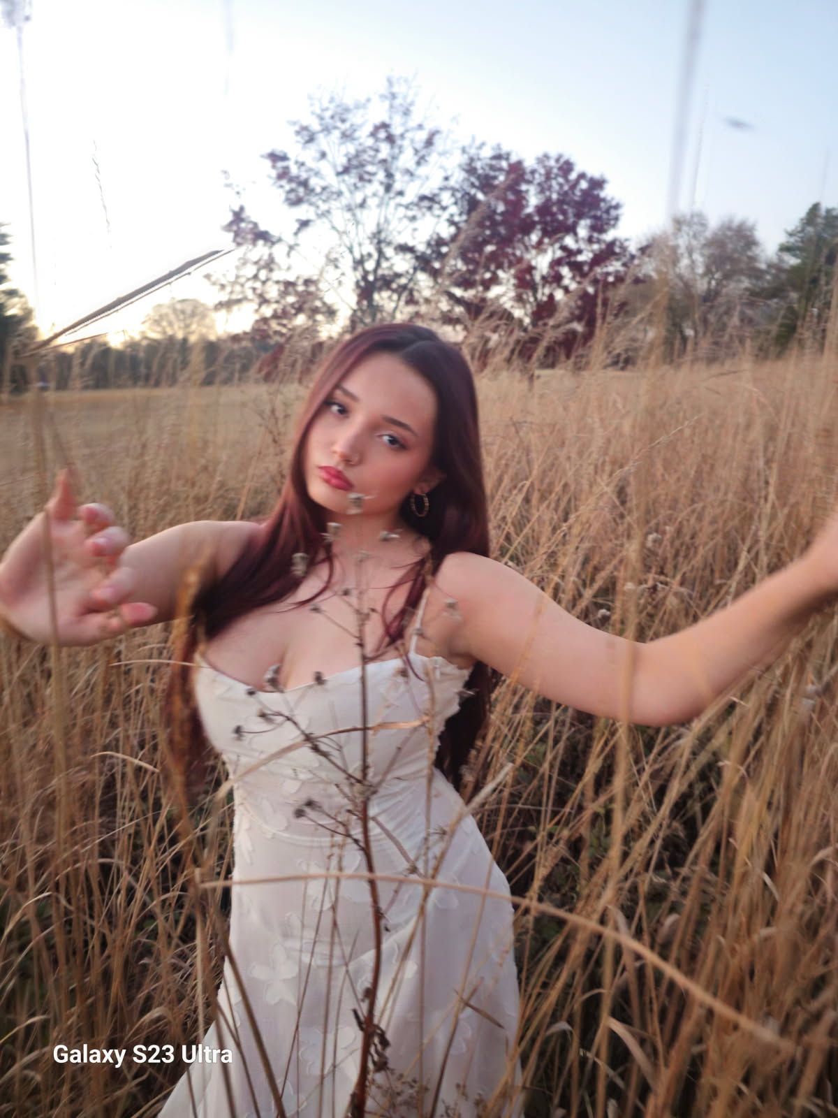Young brunette woman in white dress posing with outstretched arms in tall golden grass field at dusk.