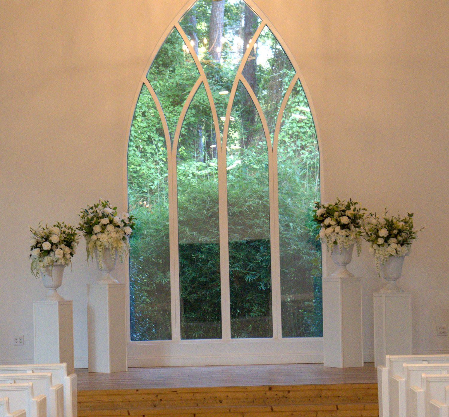 Elegant wedding chapel with Gothic arch window, white pews, and floral arrangements on pedestals overlooking lush greenery.