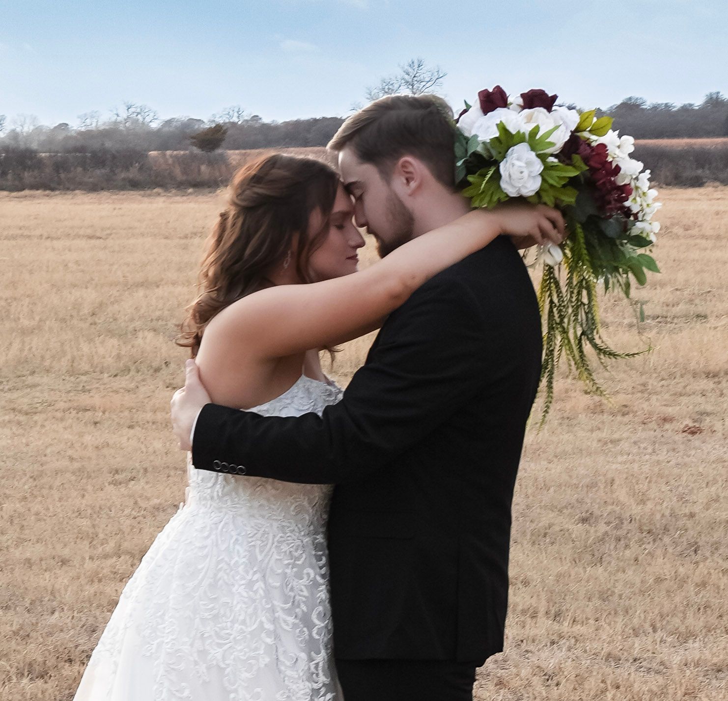 Bride and groom sharing a romantic moment in an open field, with the bride holding a red and white bouquet.