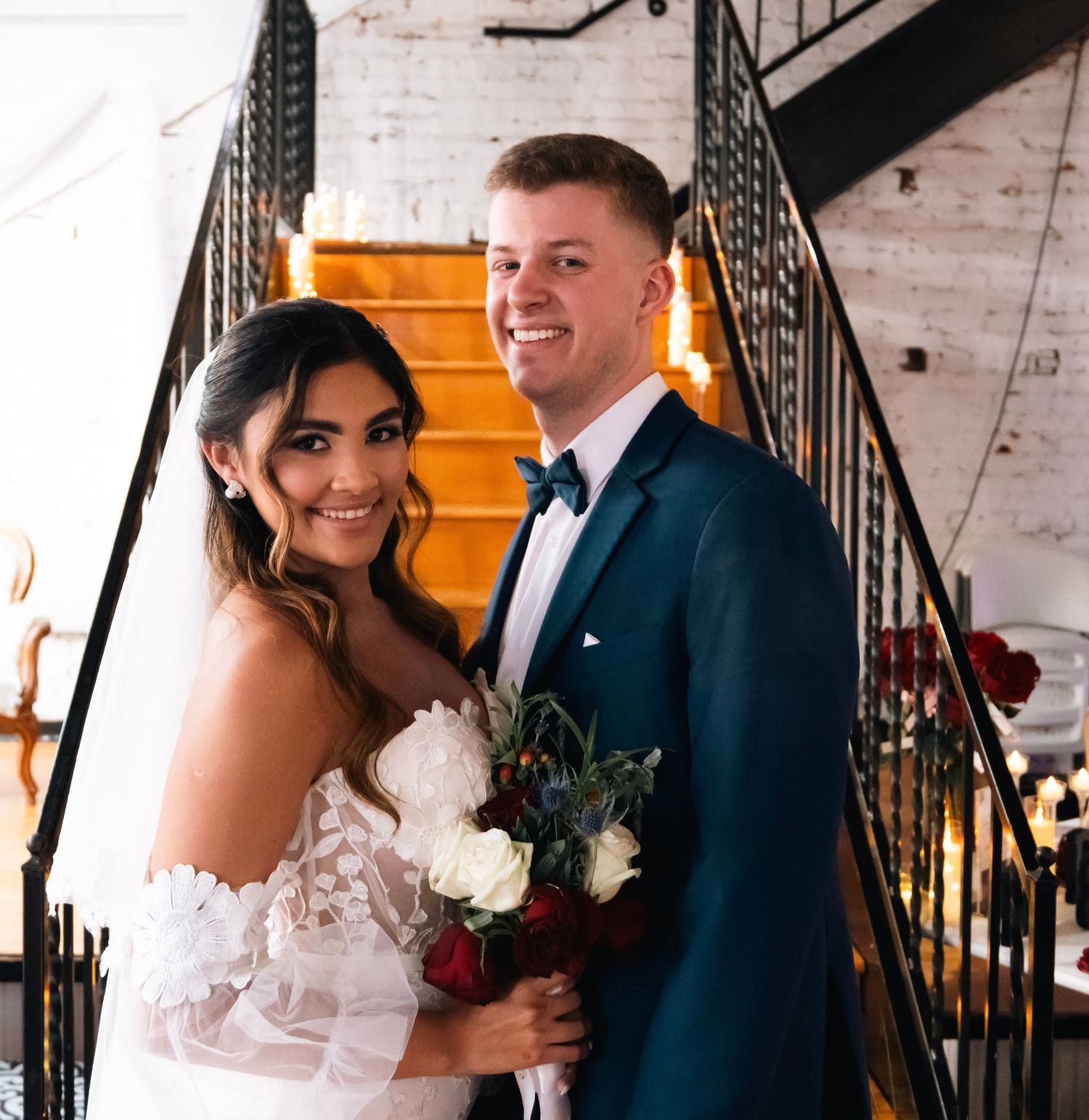 Smiling bride and groom posing on staircase, she in off-shoulder lace gown, he in navy tuxedo.