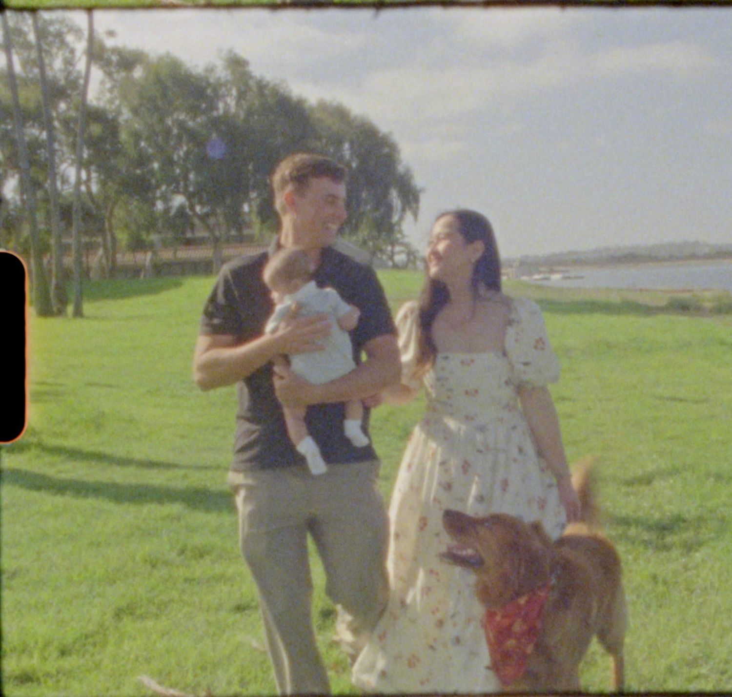 Smiling couple carrying baby with golden retriever on sunny green lawn near water.