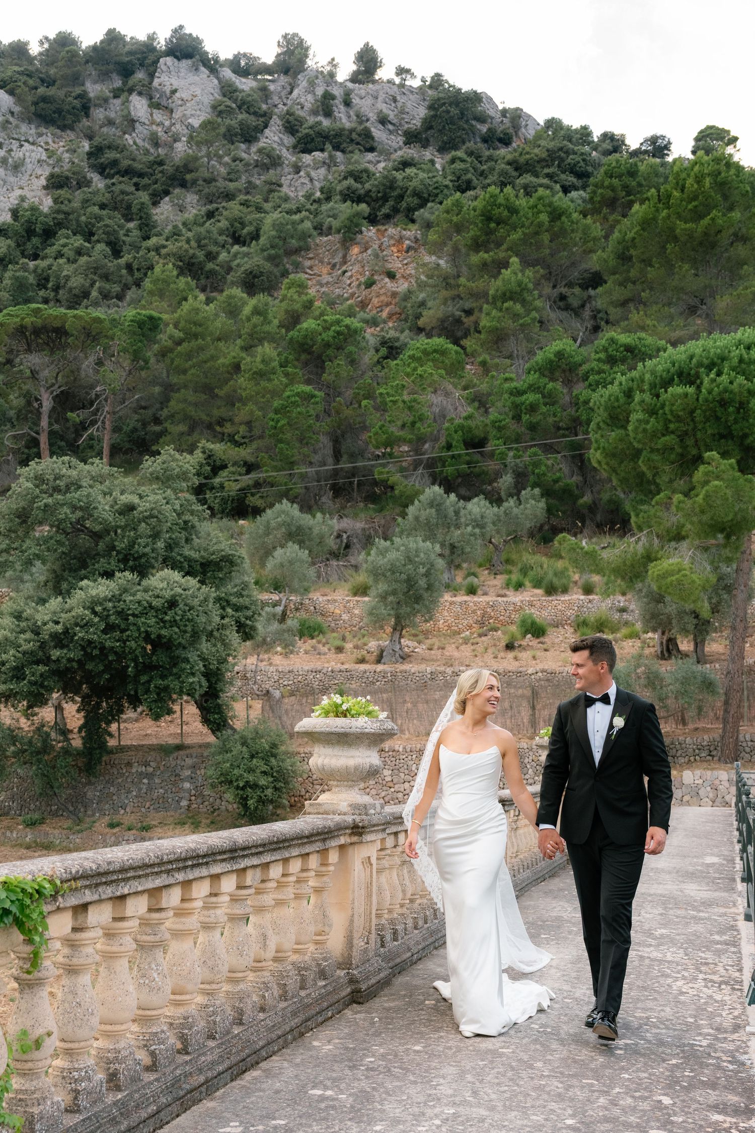 Bride and groom walk hand-in-hand along a stone balustrade terrace with lush green hillside backdrop.