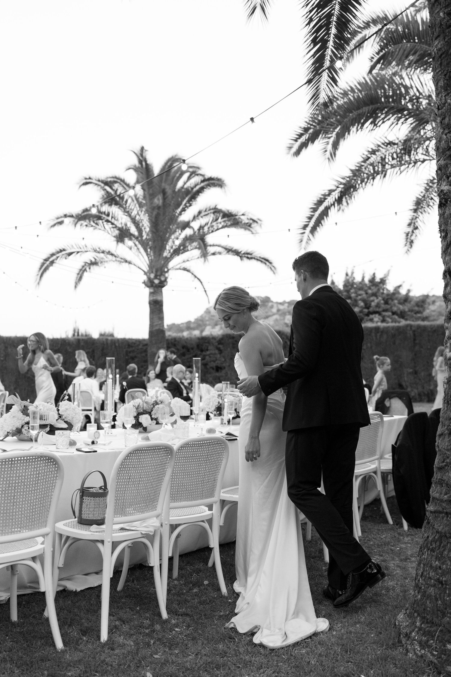 Black and white photo of bride and groom walking together at outdoor wedding reception with palm trees and elegant table setting.