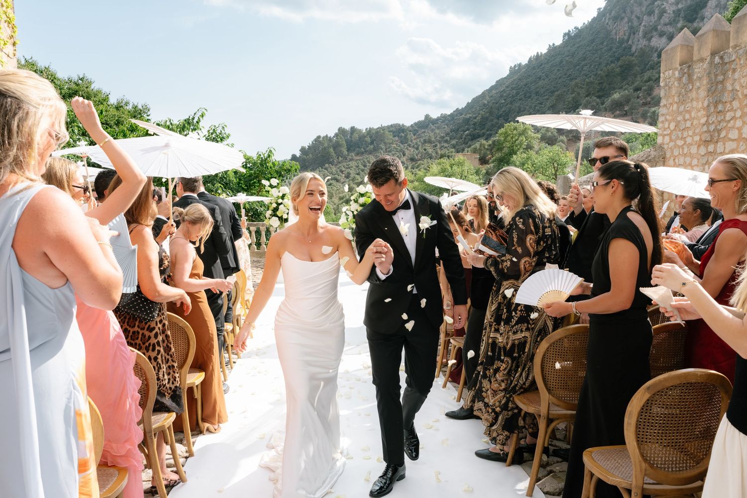 Bride and groom walk down outdoor aisle as guests celebrate with flower petals at a scenic Mediterranean wedding.