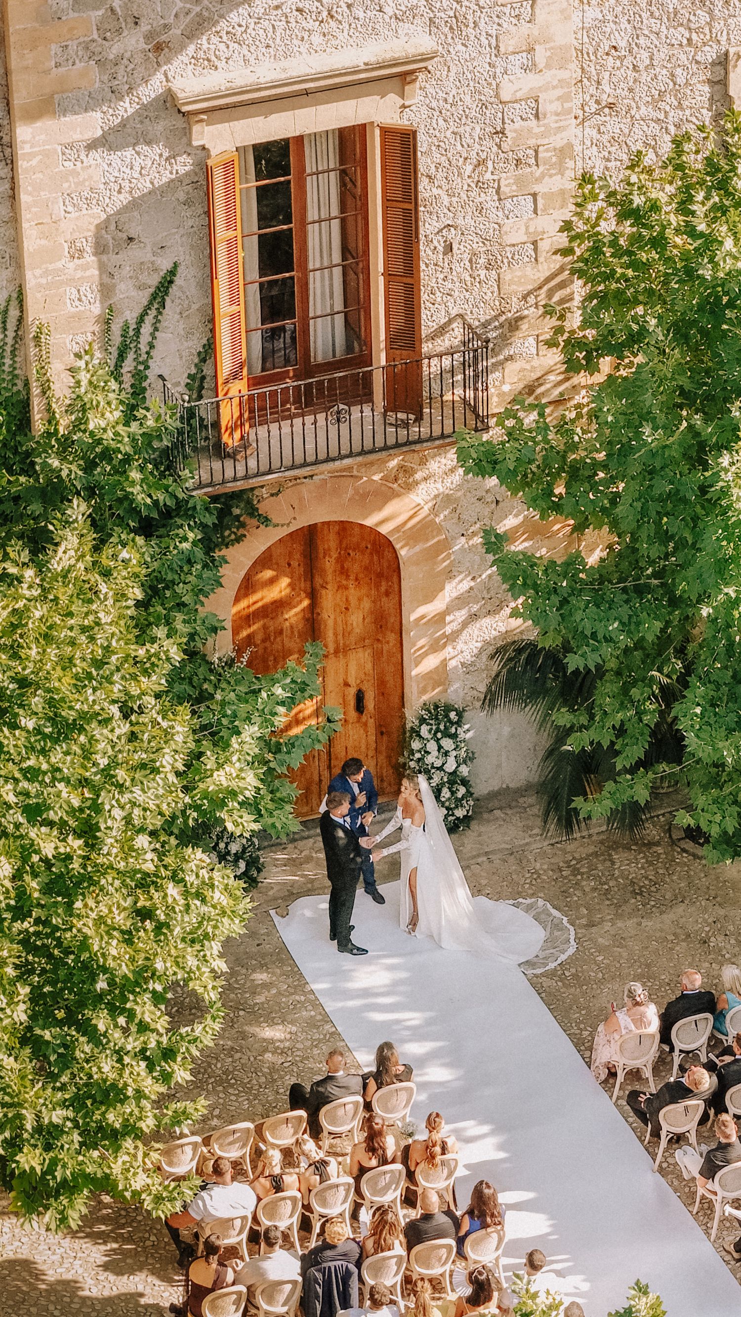 Aerial view of an outdoor wedding ceremony at a rustic stone villa with lush greenery and guests seated along a white aisle.