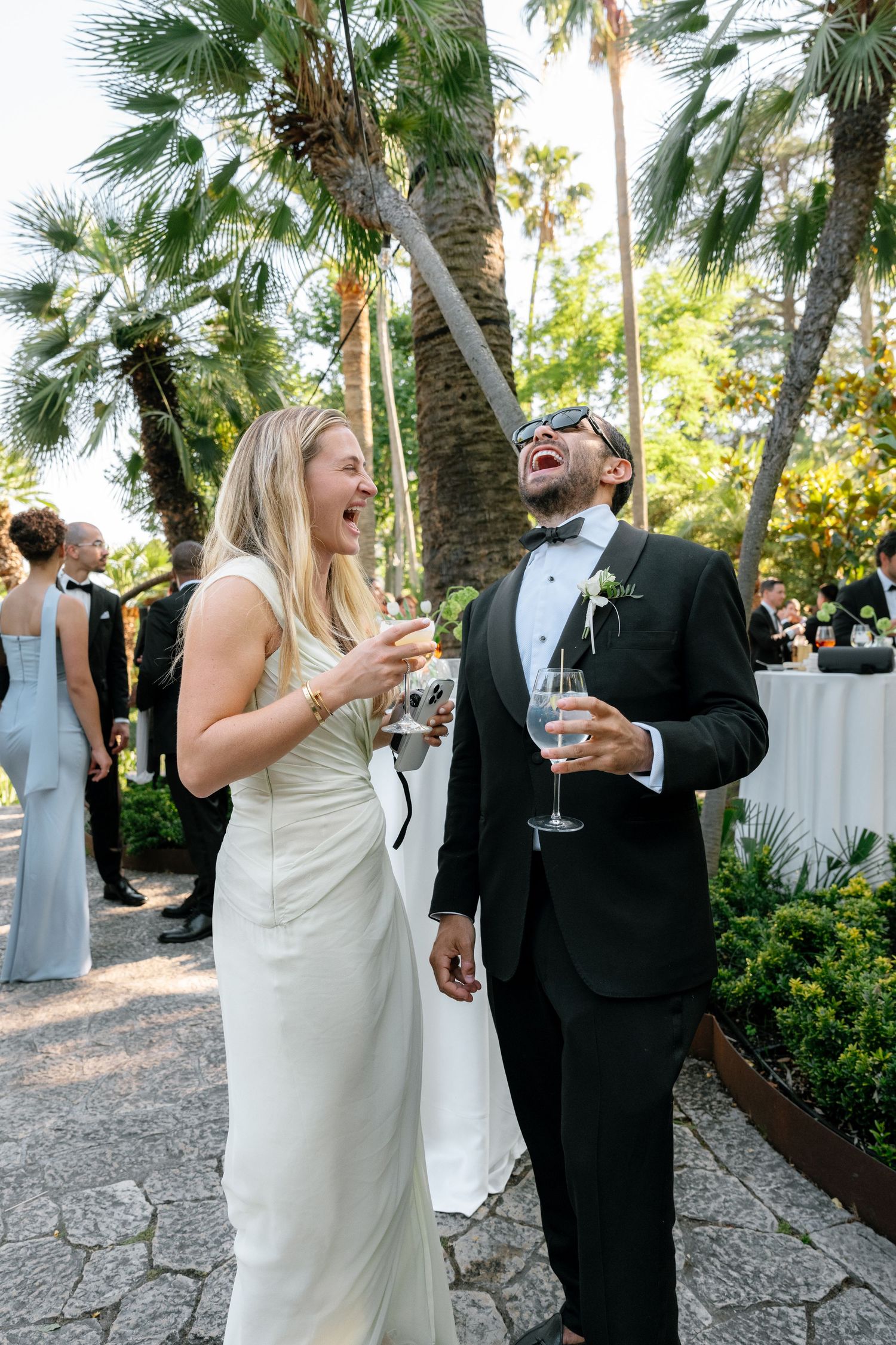 Bride and groom laughing joyfully outdoors at wedding reception, holding champagne glasses among palm trees.