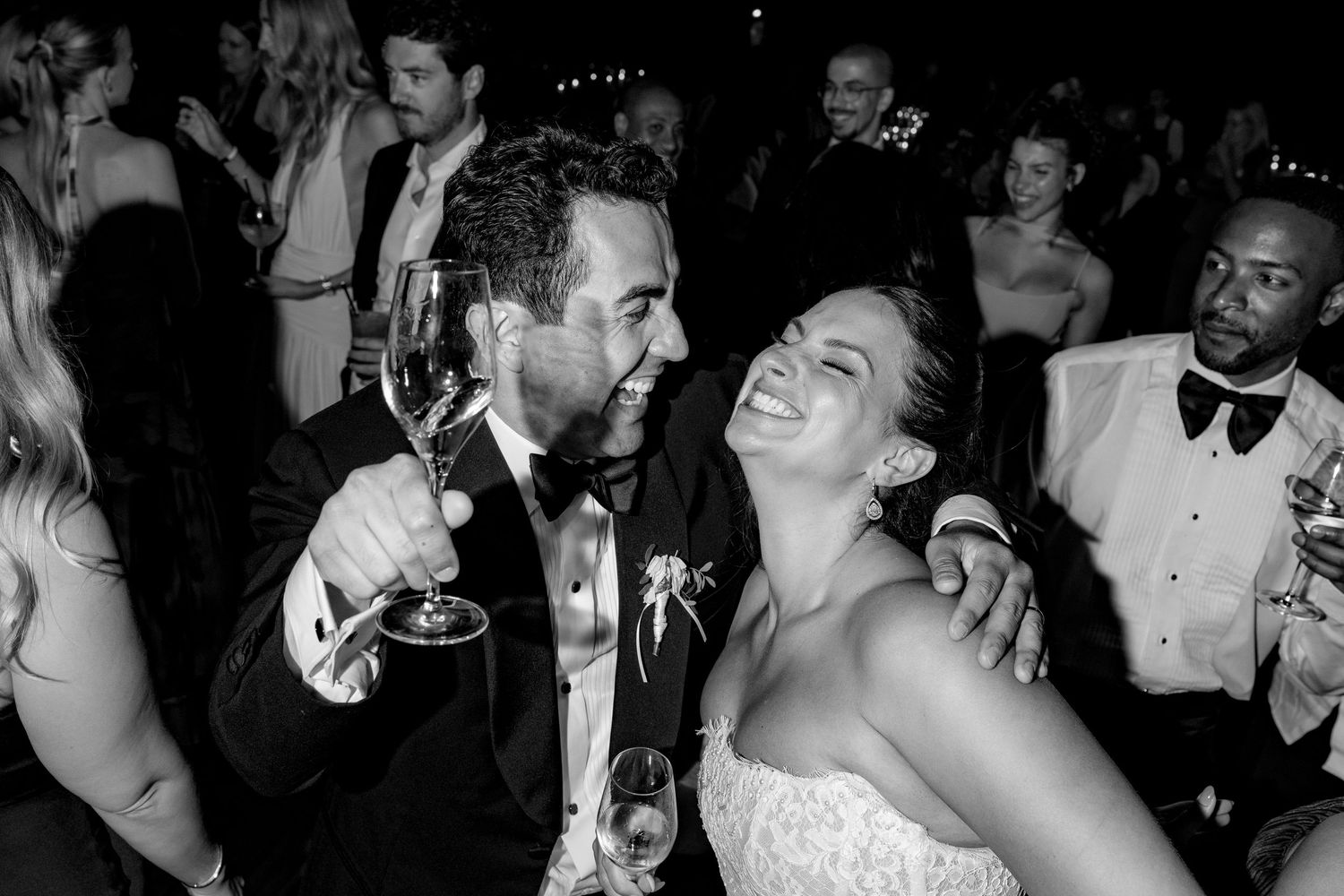 Black and white photo of a laughing bride and groom toasting with champagne glasses at their wedding reception.