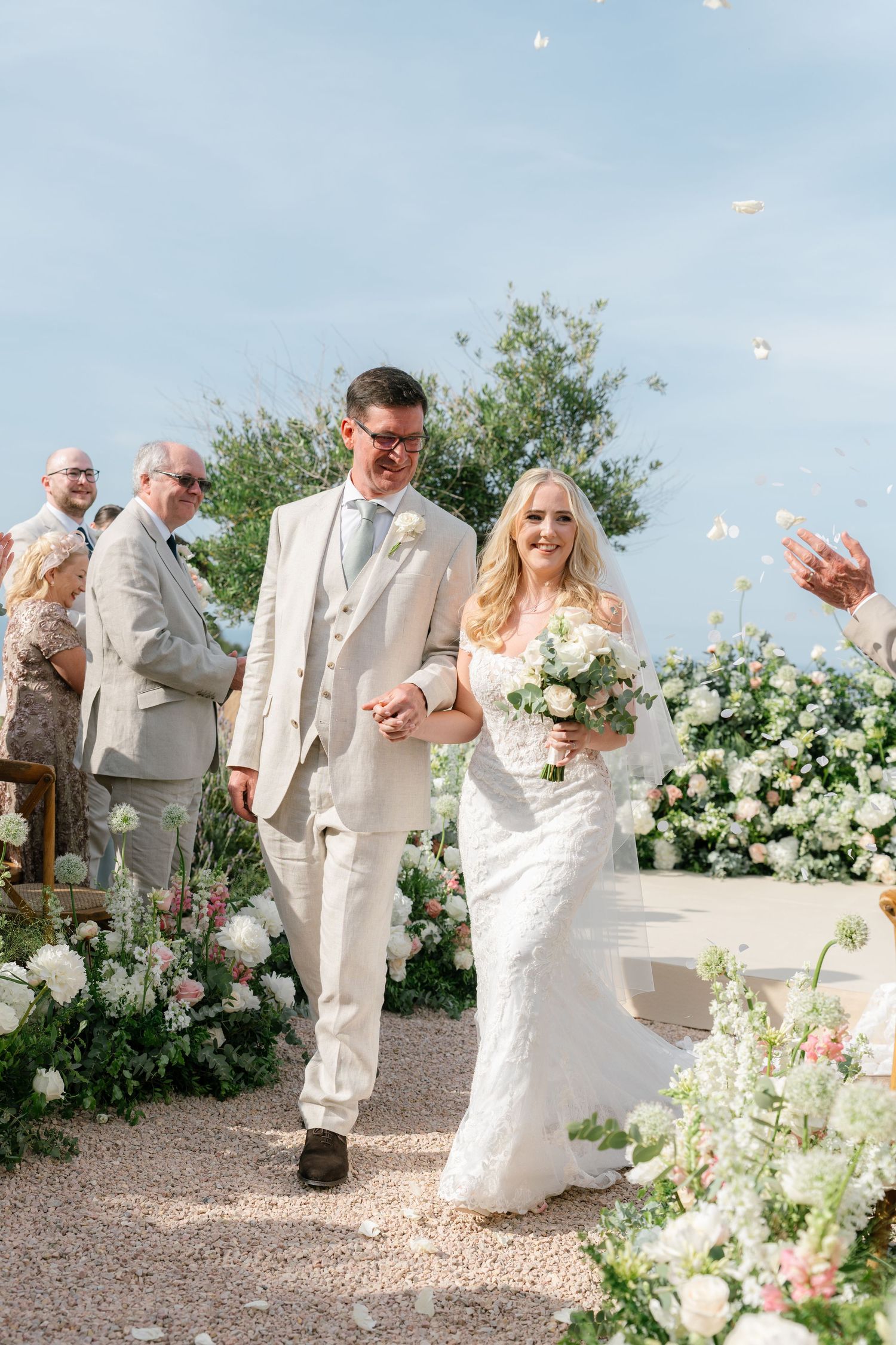 Newlyweds celebrating with confetti toss in light colored attire against floral backdrop.