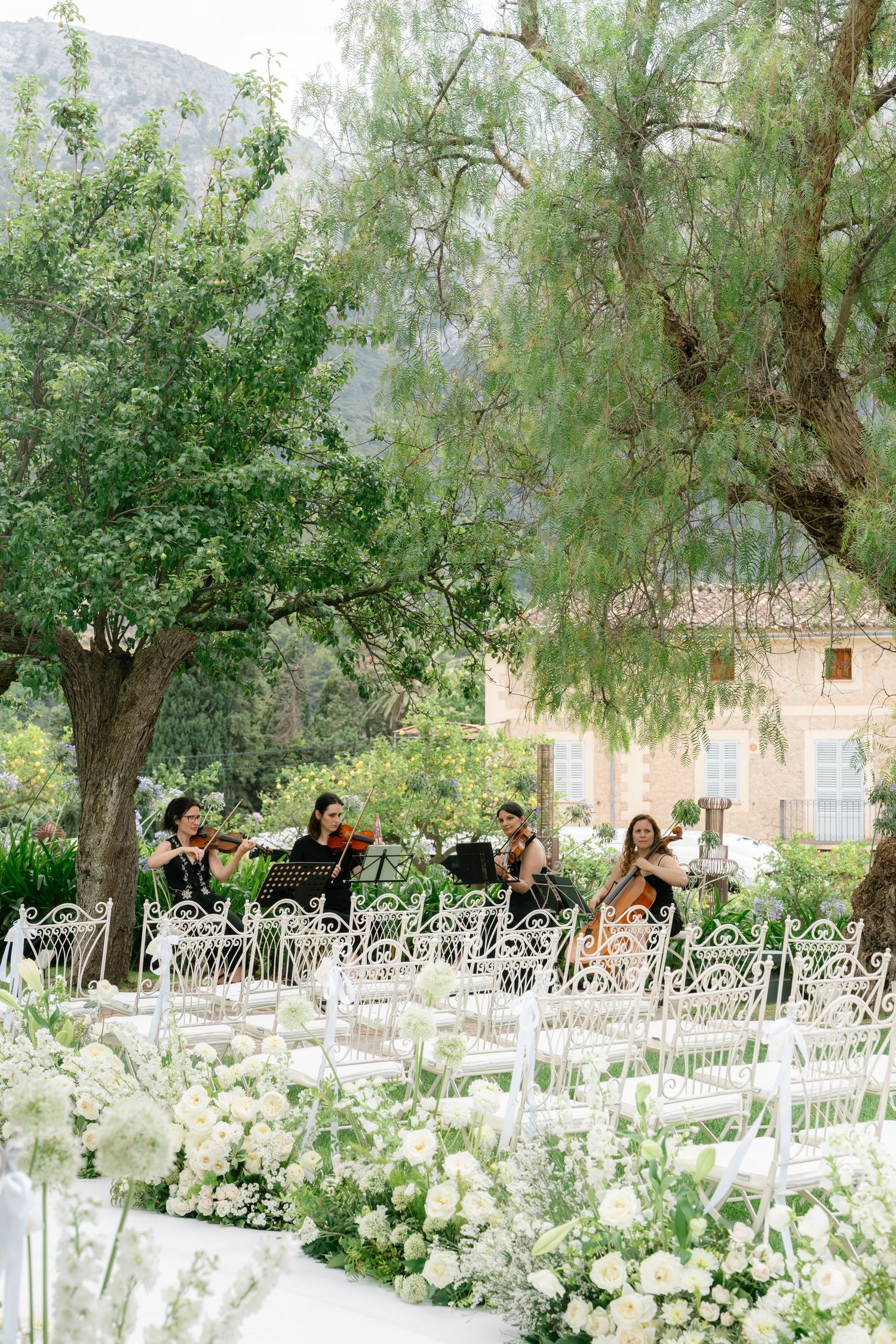 Garden wedding ceremony setup with white chairs and floral arrangements under shady trees.
