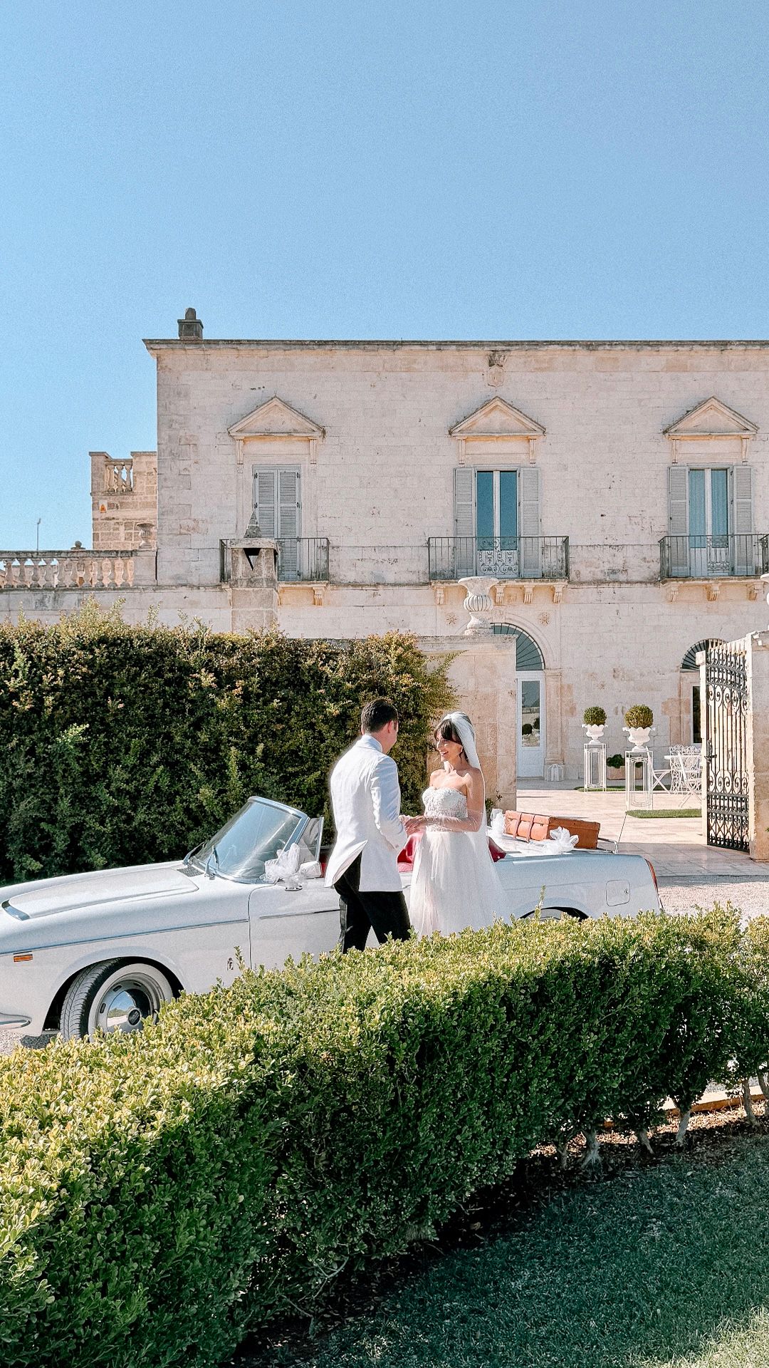 Elegant wedding scene with classic cars and white stone buildings against a green hedge backdrop.