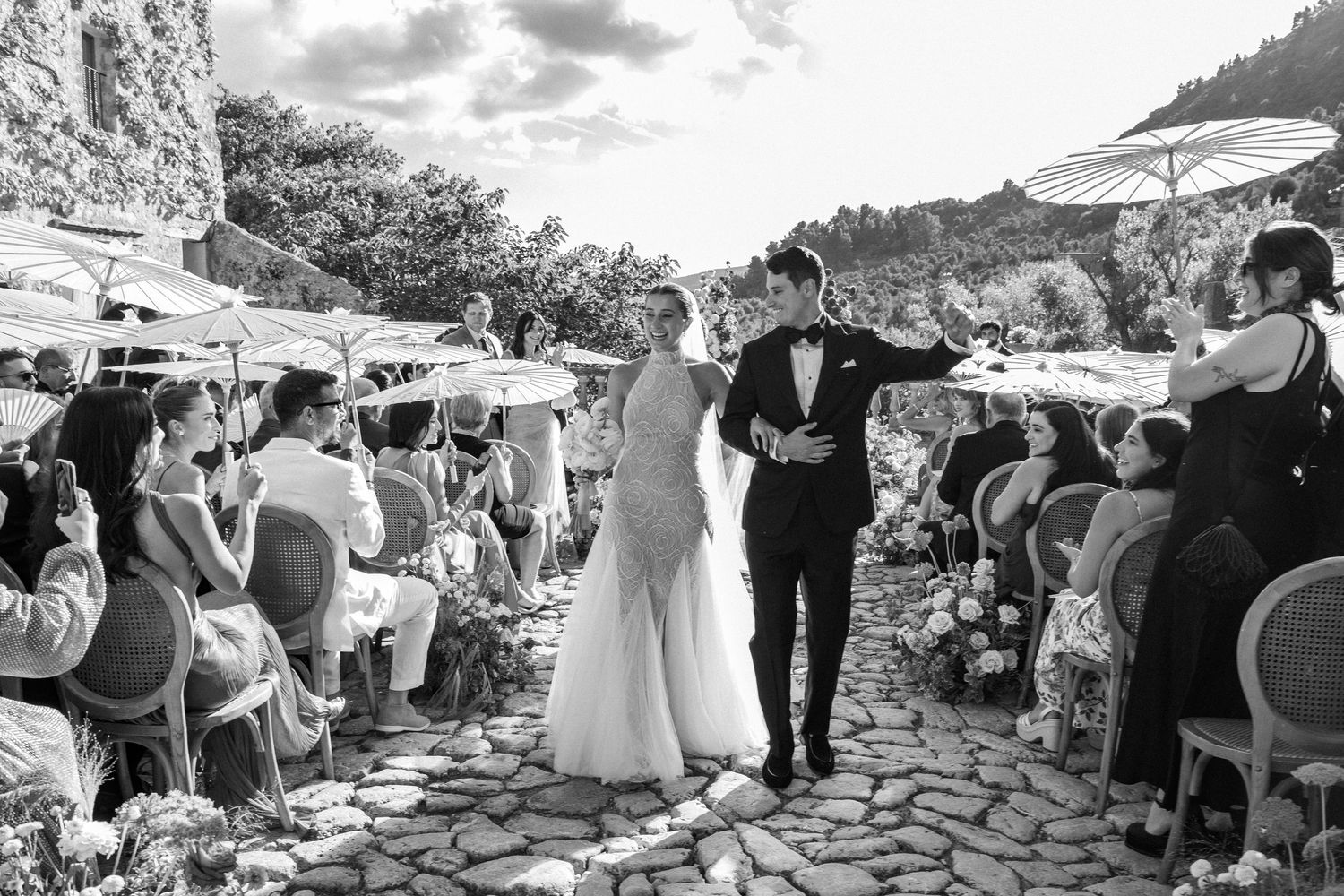 Wedding ceremony guests seated outdoors under white umbrellas on stone patio.