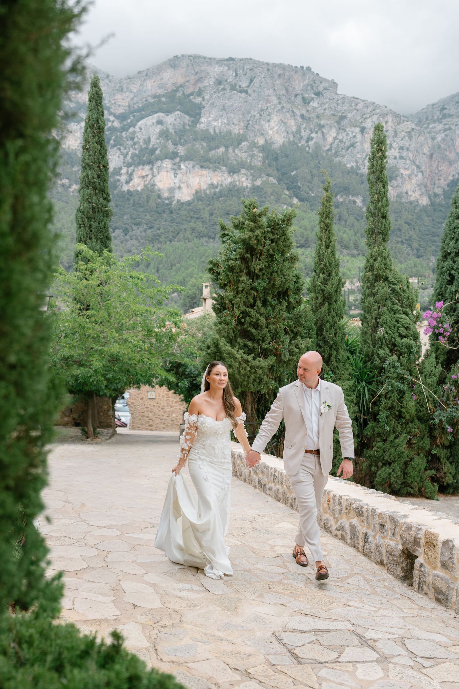 Wedding couple walking along a stone pathway with cypress trees and mountains in the background.