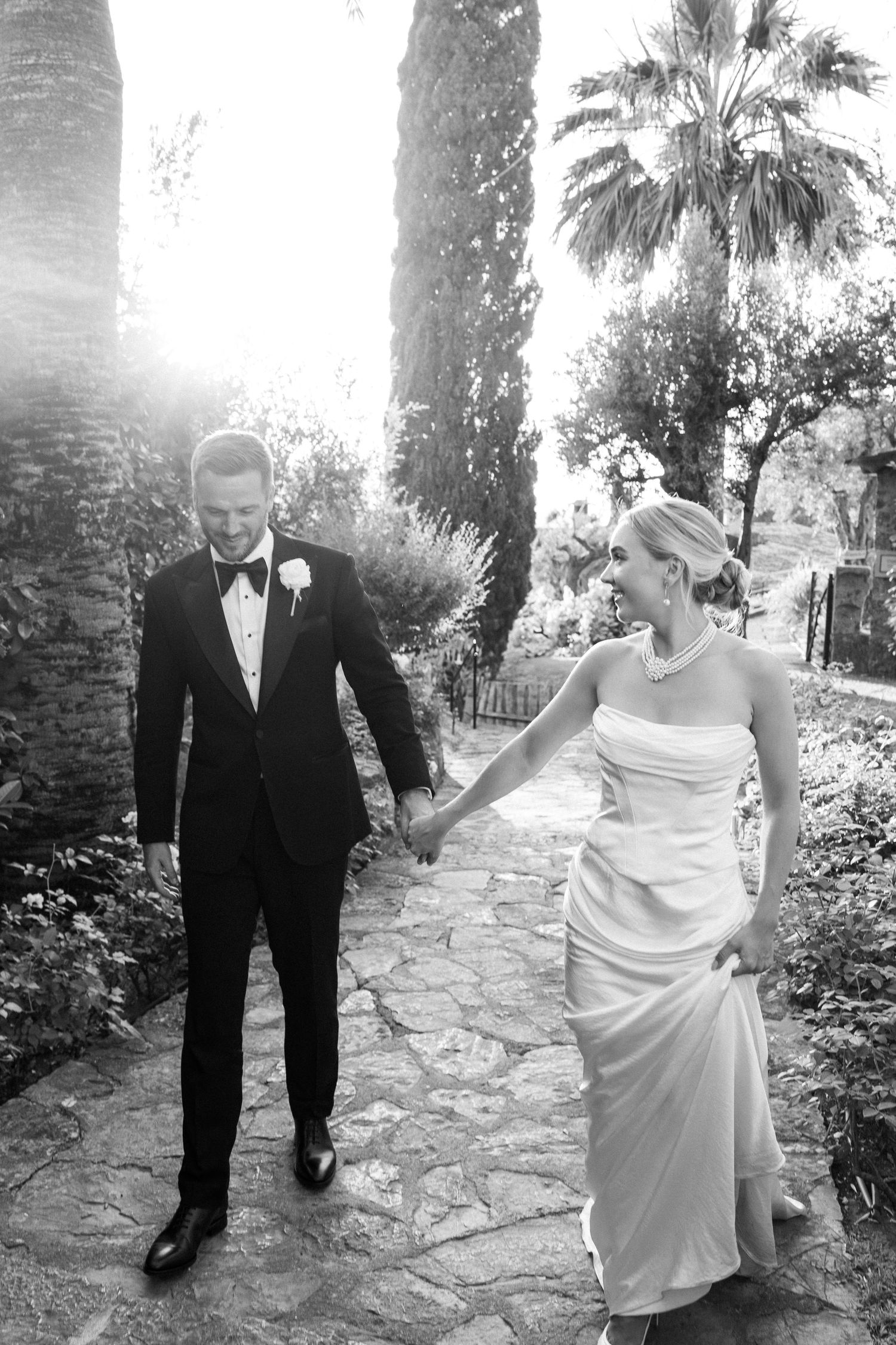Wedding couple shares a playful moment on a stone path lined with palm trees.