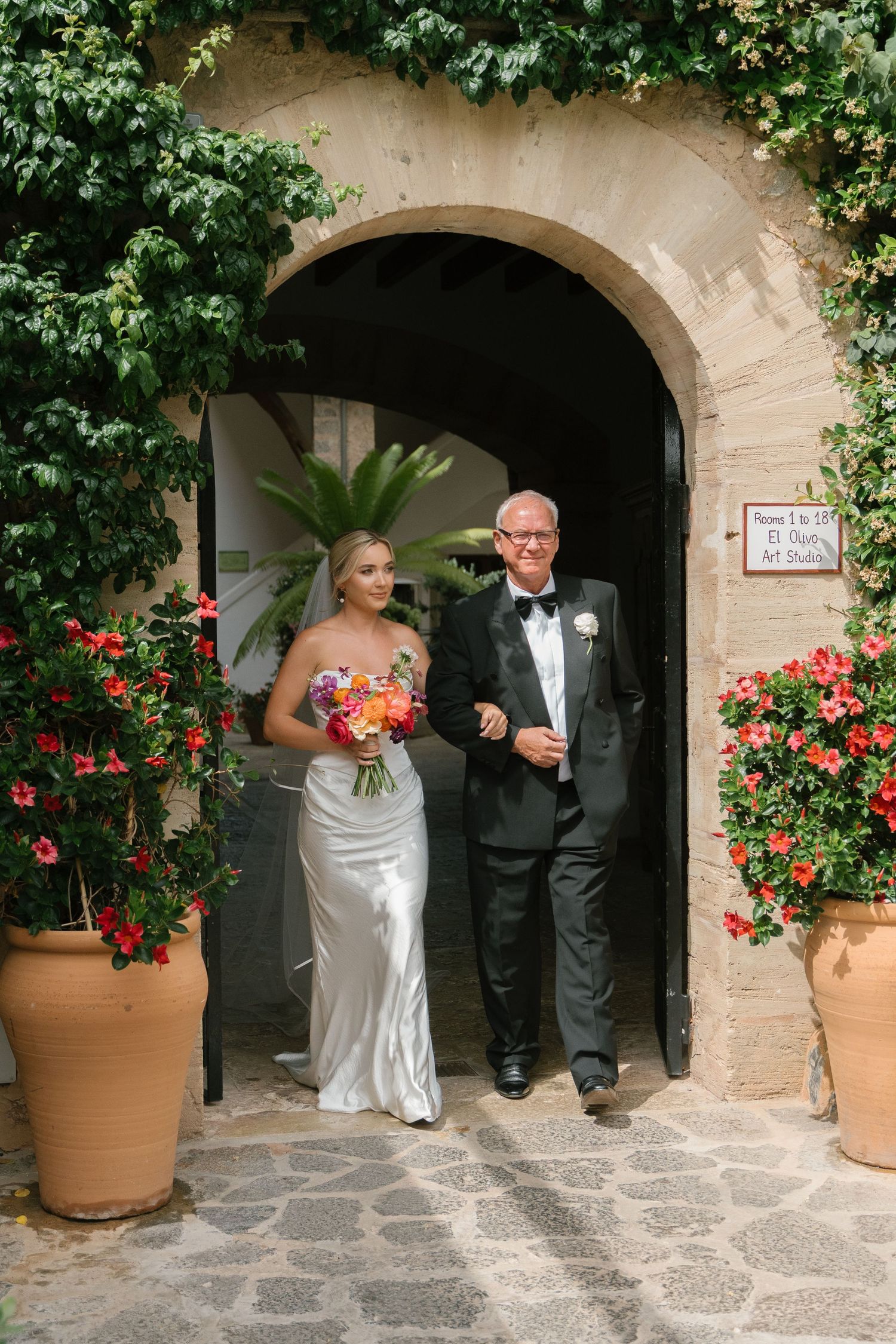Wedding entrance through stone archway decorated with vibrant red flowers in terracotta pots.