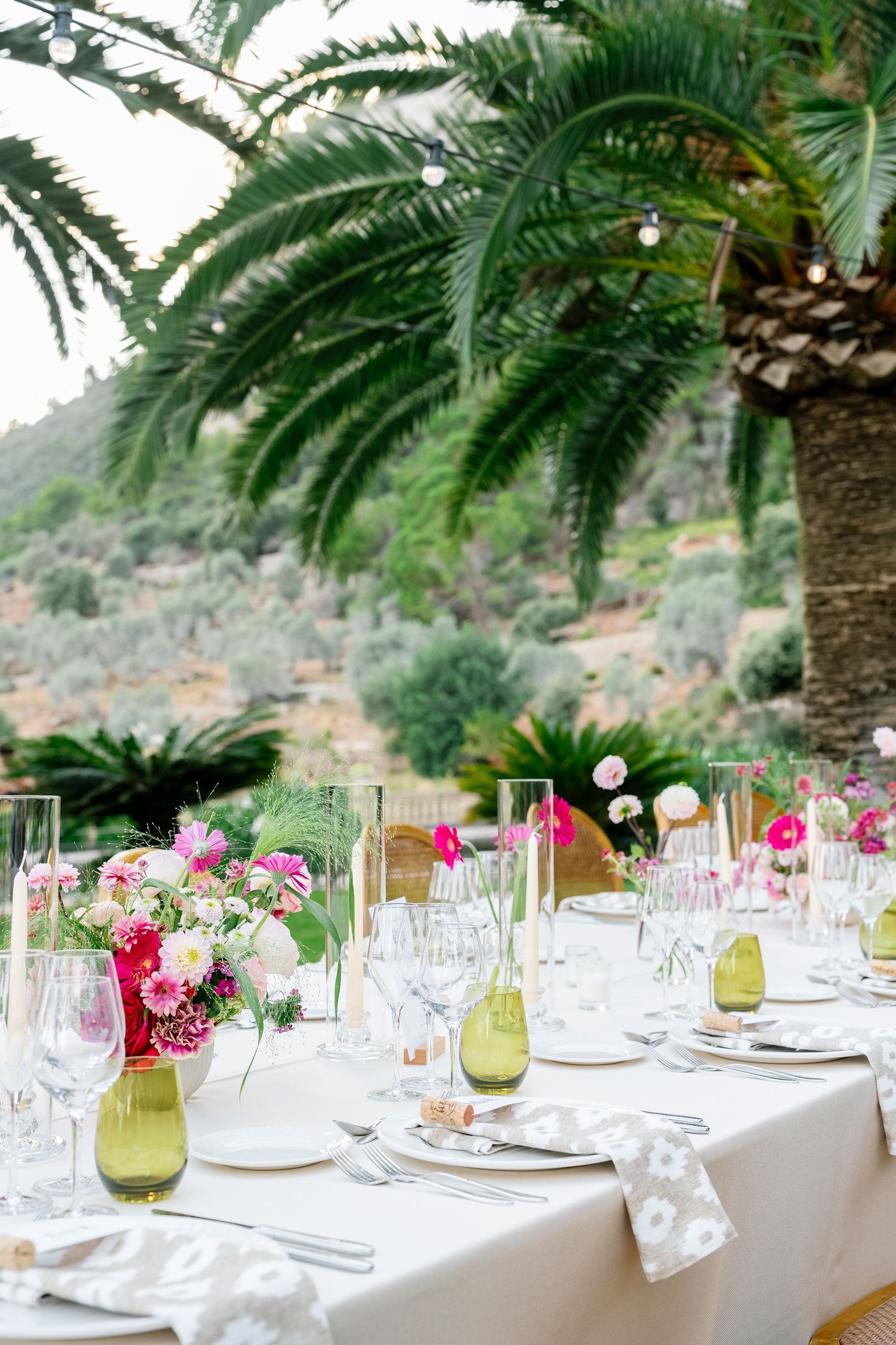 Elegant outdoor wedding reception table setting with palm trees and pink floral centerpieces.