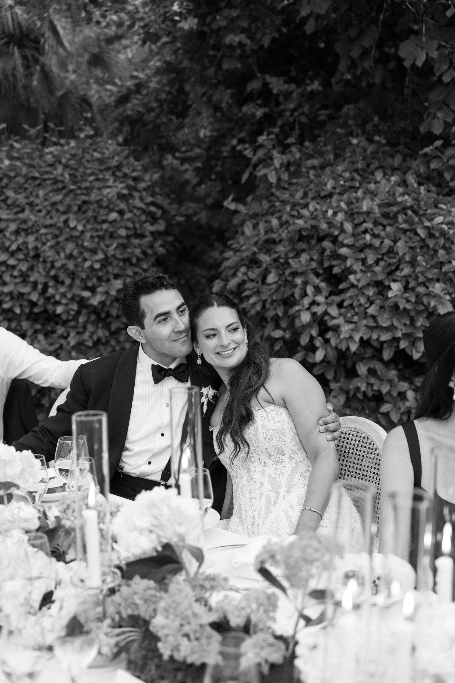 Black and white photo of a smiling bride and groom seated at an elegant outdoor wedding reception table.