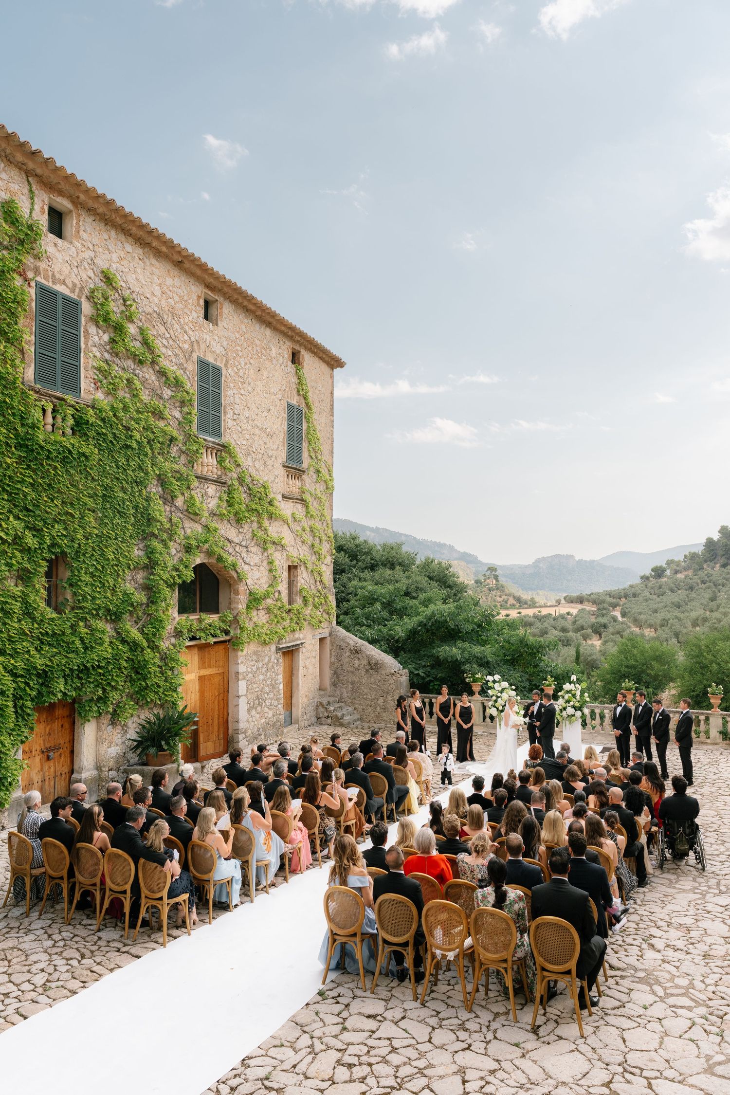Outdoor wedding ceremony at a rustic stone villa covered in ivy, with guests seated on wooden chairs overlooking scenic mountains.