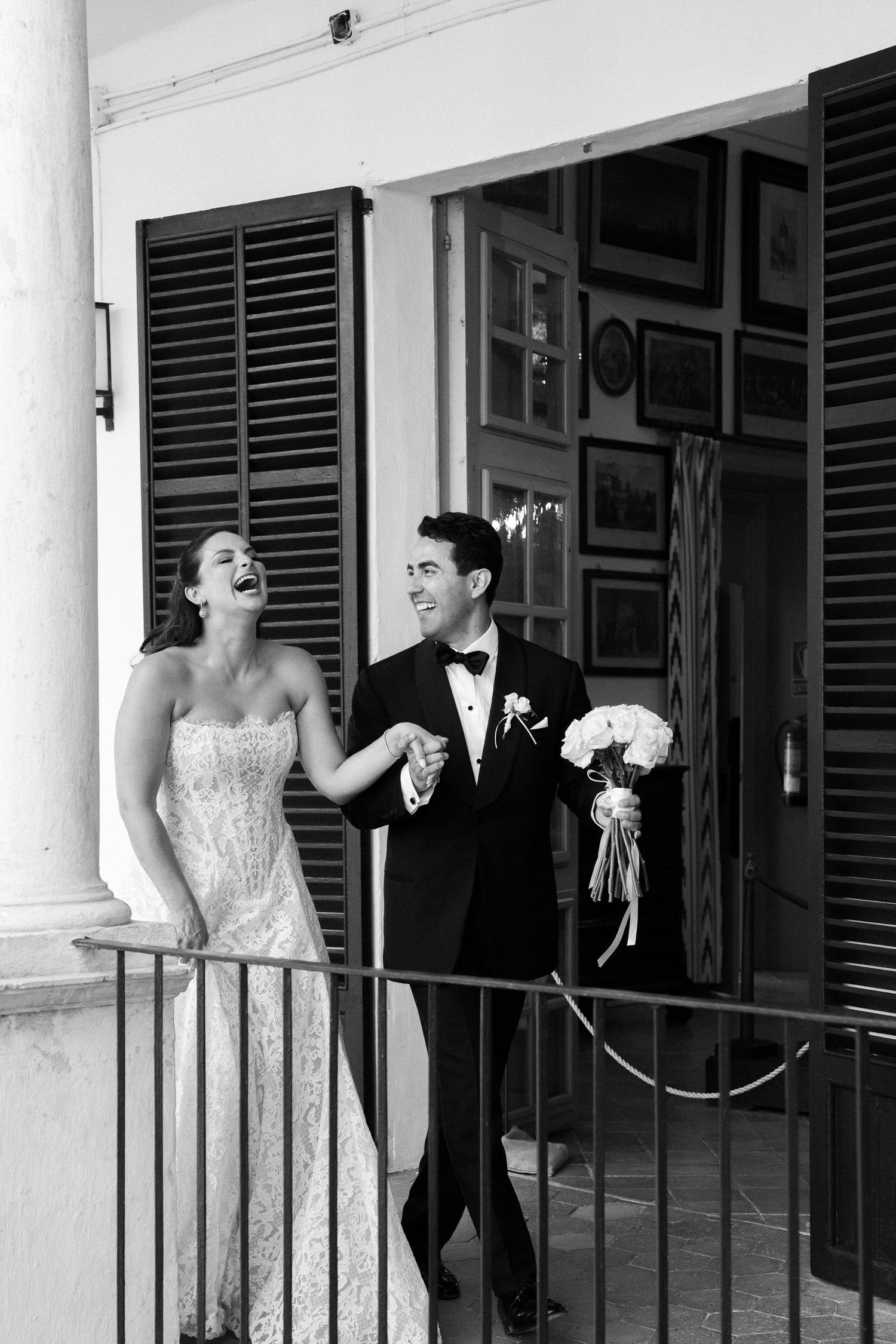 Happy bride and groom laughing on a balcony, bride in lace gown, groom in tuxedo holding bouquet, black and white wedding photo.