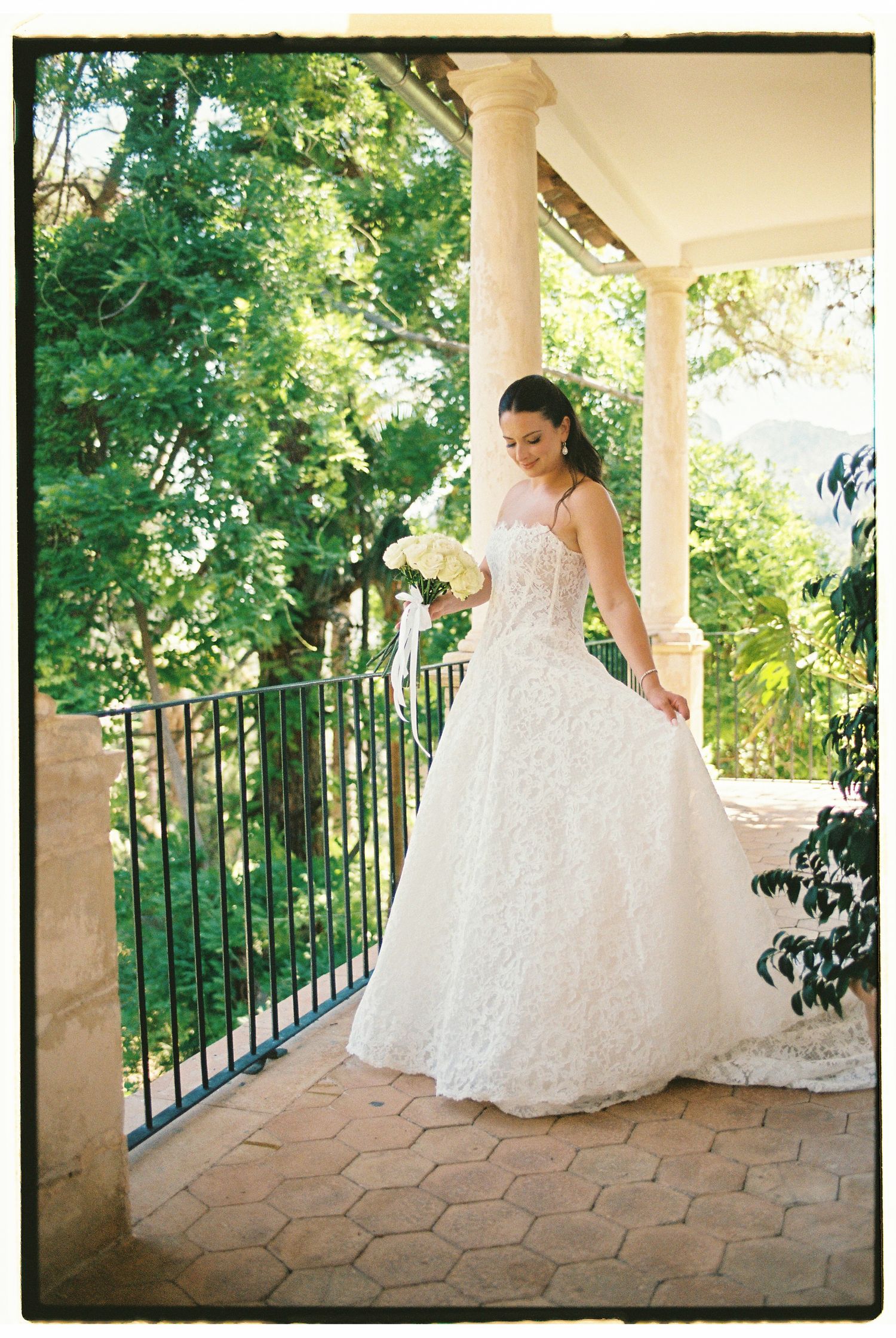 Bride in white lace ball gown holding bouquet on elegant stone balcony surrounded by lush green trees.