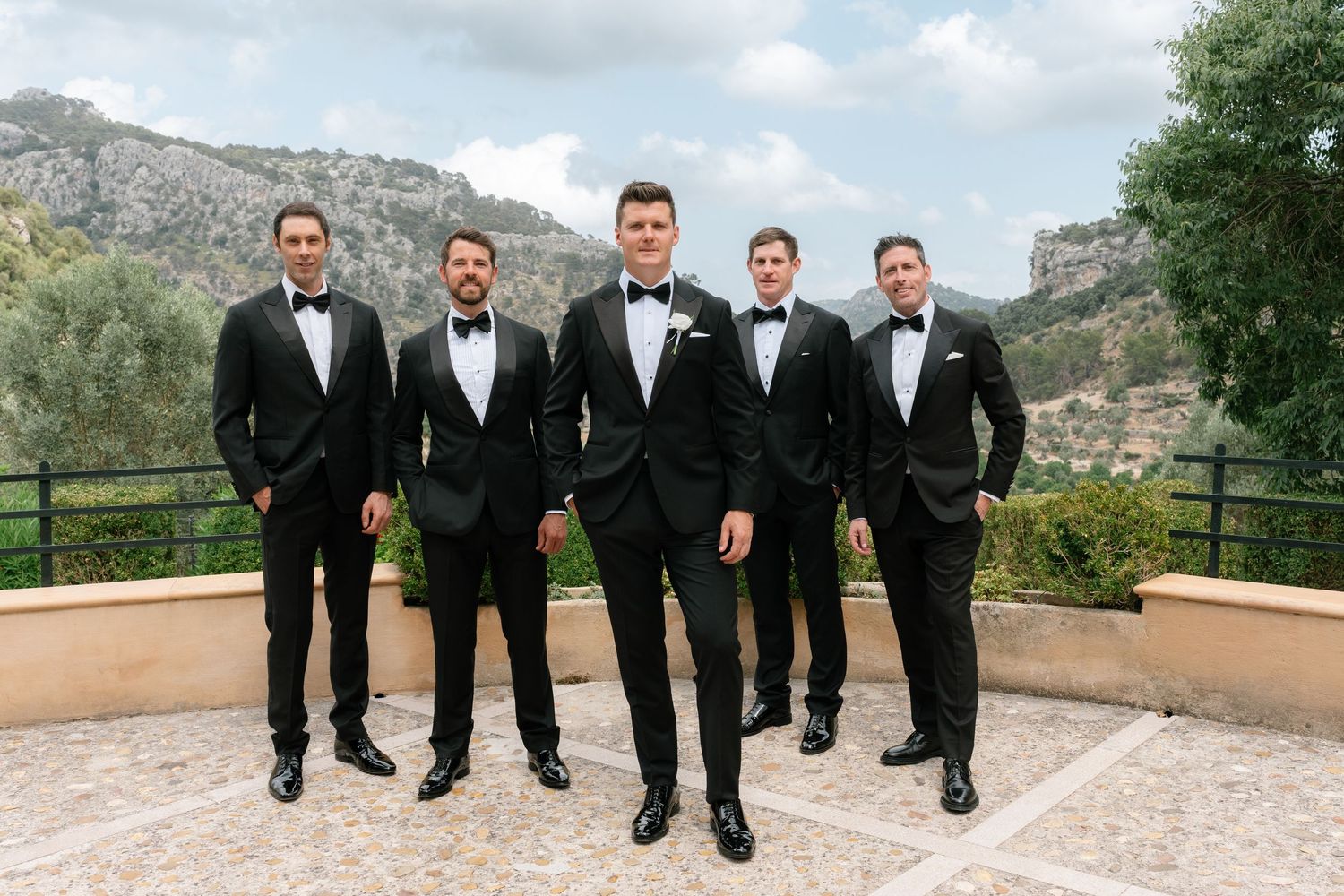 Five groomsmen in black tuxedos and bow ties pose on an outdoor terrace with mountain scenery behind them.
