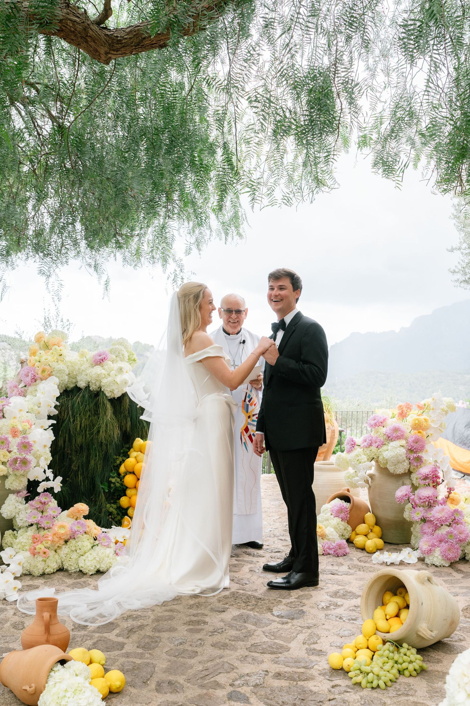 A wedding couple poses on a beach with colorful floral arrangements and willow trees creating a romantic setting.