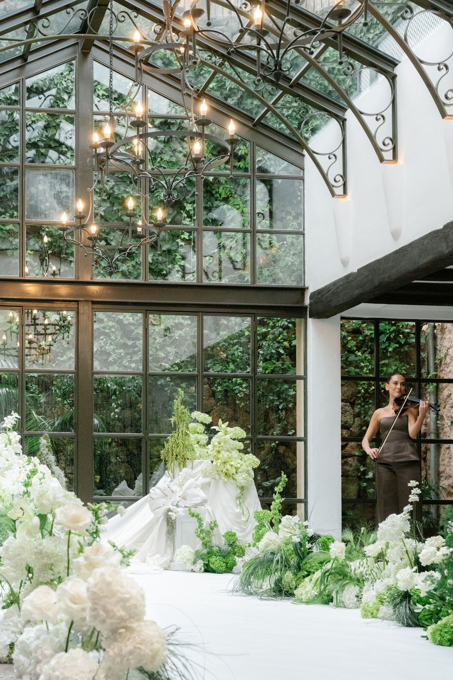A sunlit greenhouse wedding venue decorated with white floral arrangements and elegant chandeliers.