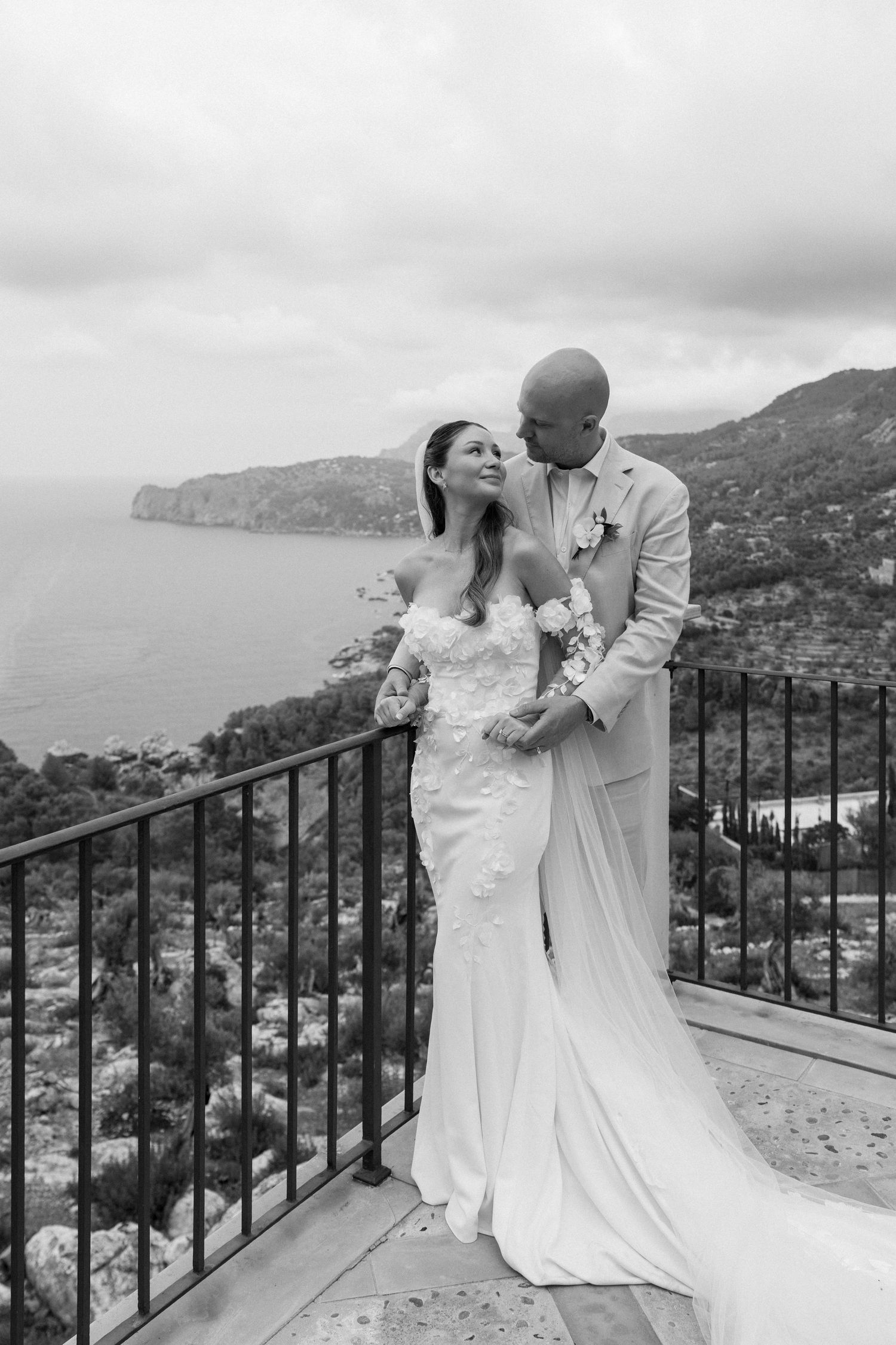Newlyweds share romantic moment on balcony overlooking Mediterranean coastline.