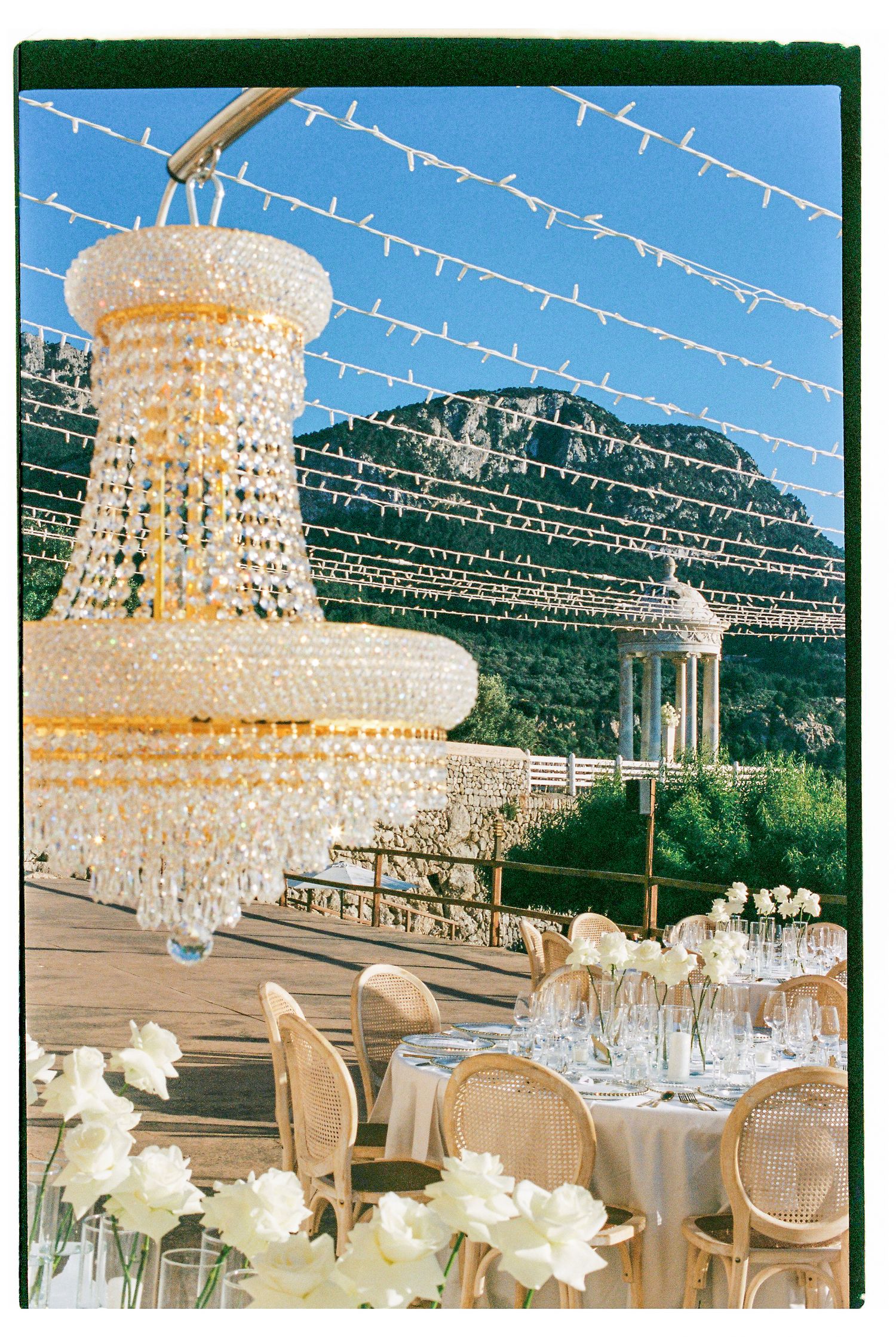 Elegant wedding reception setup with crystal chandeliers and string lights against mountain backdrop.