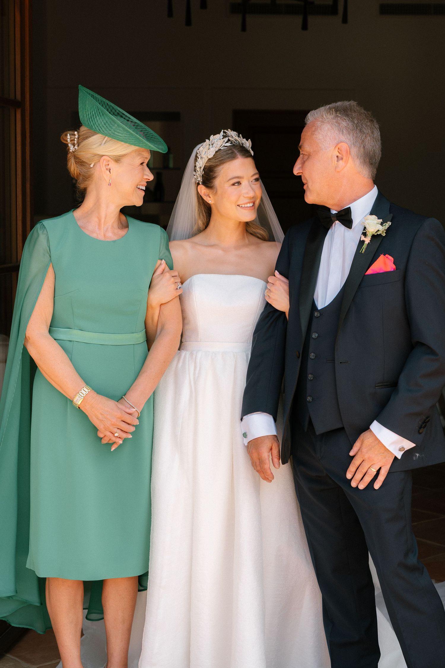 Wedding party poses in elegant green dresses and formal attire against dark interior.