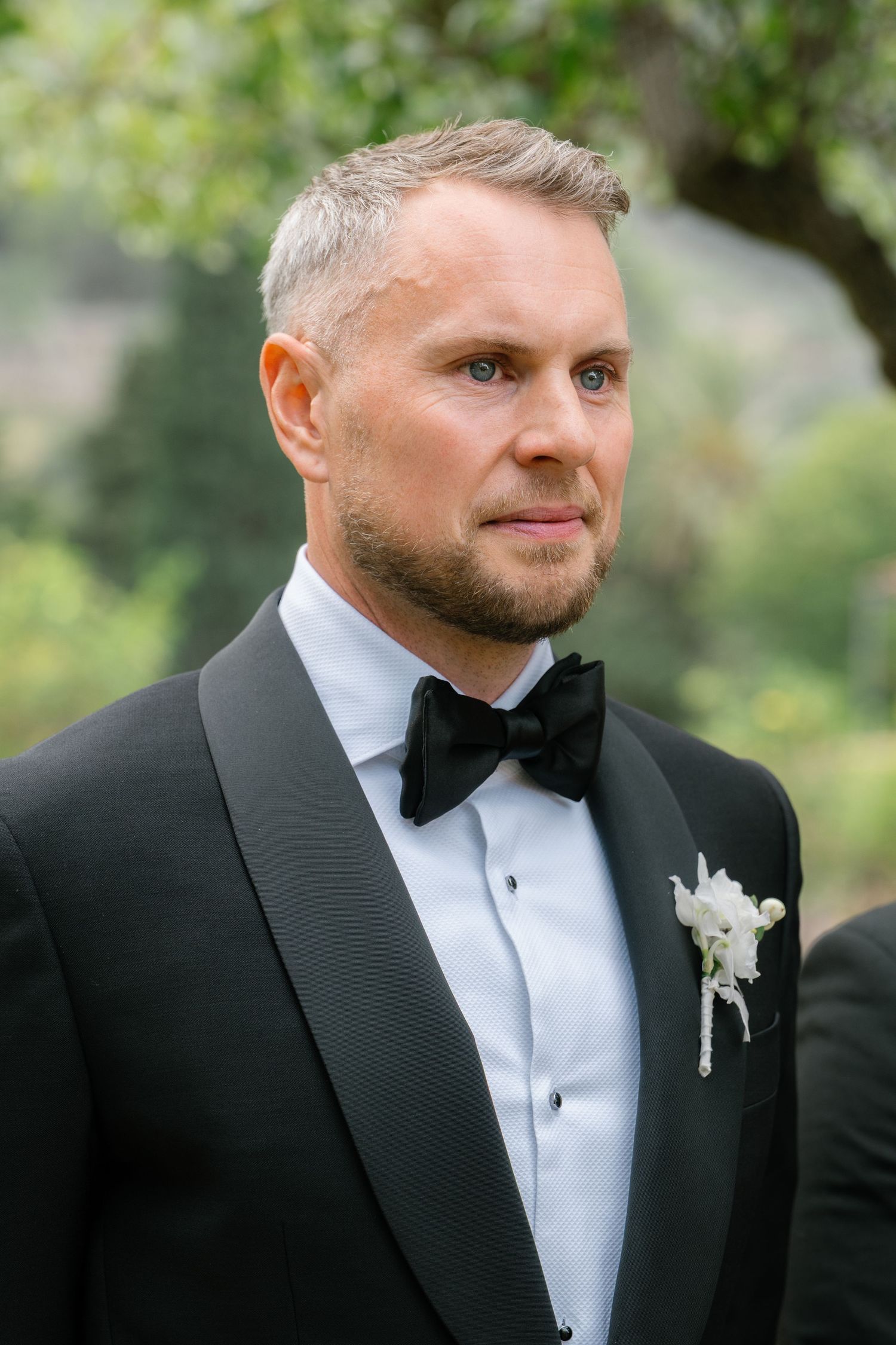 Groom in classic black tuxedo and white boutonniere at outdoor wedding.
