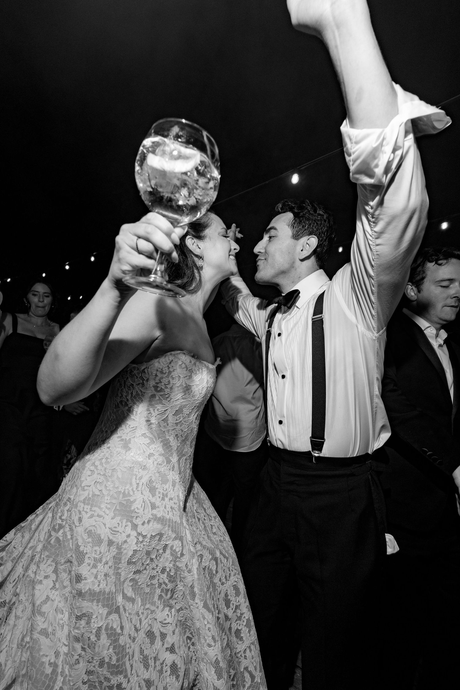 Black and white photo of a bride and groom celebrating at their wedding reception, dancing joyfully with a large cocktail glass.