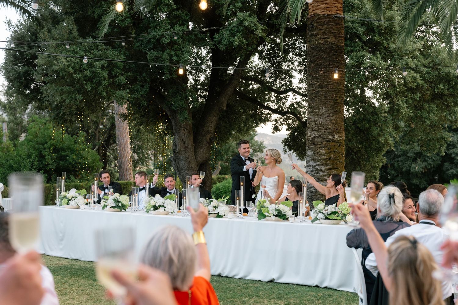 Outdoor wedding reception toast with bride and groom at head table surrounded by lush greenery and string lights.