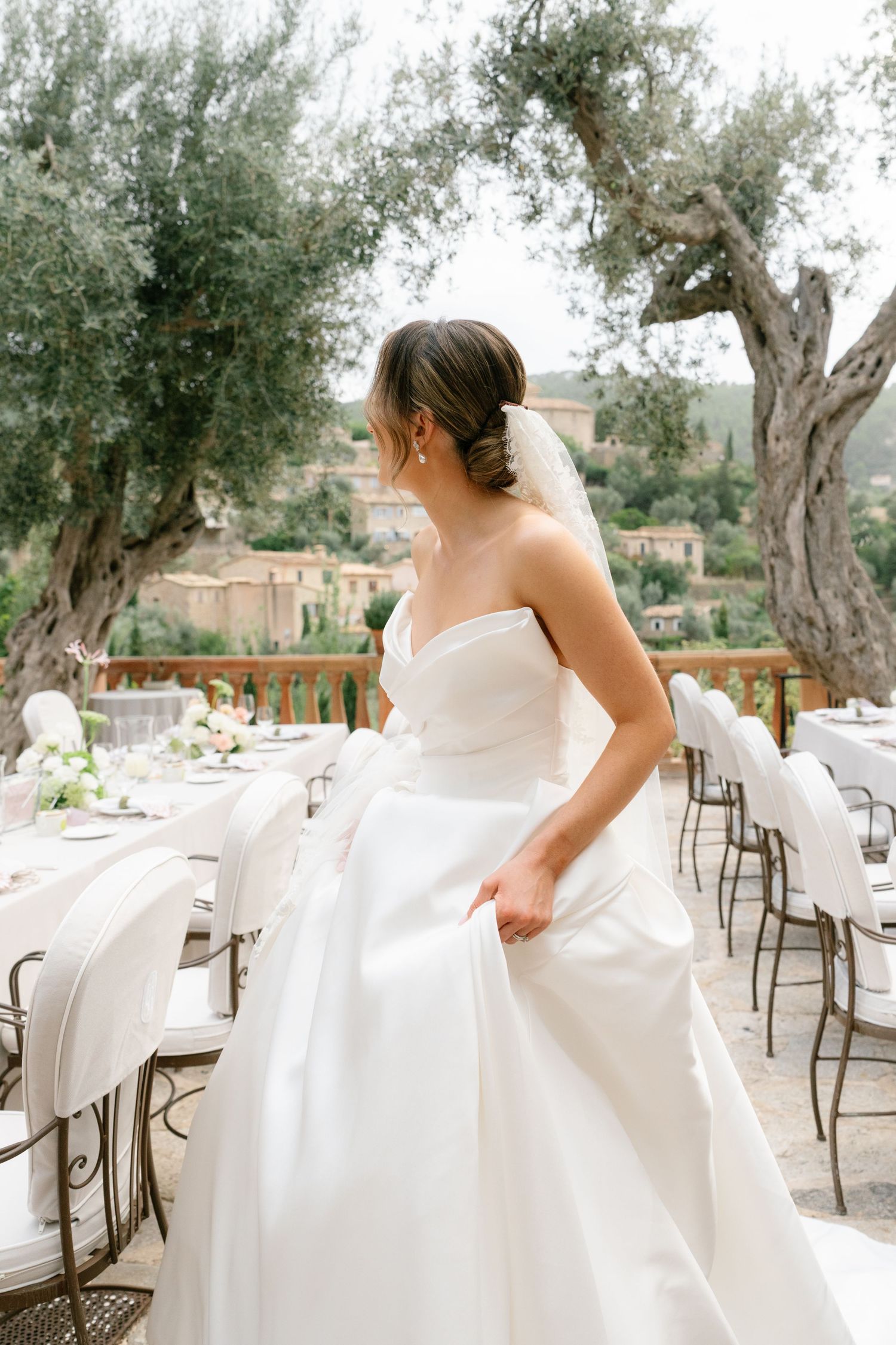 Bride in flowing white dress walking through an outdoor terrace with olive trees.