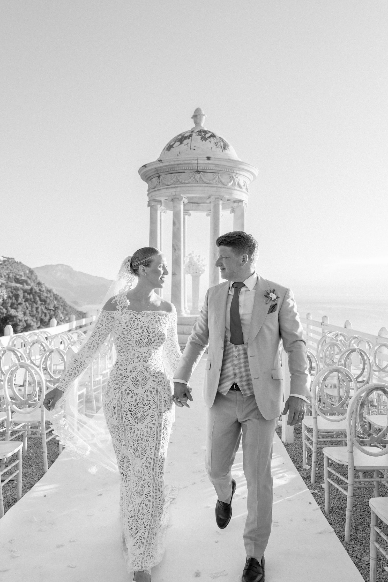 Joyful wedding couple walking through classical pavilion with ornate white chairs.