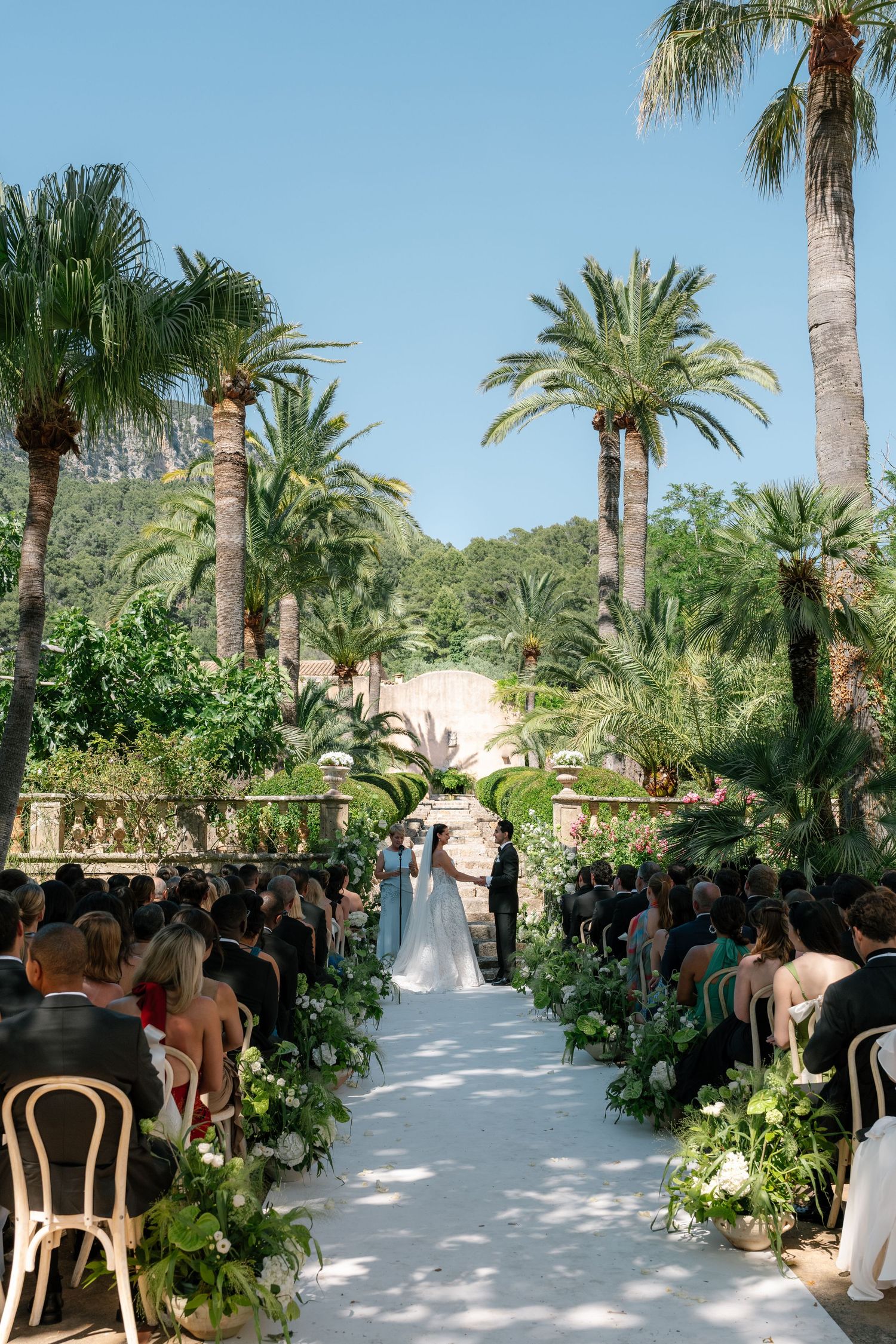 Outdoor wedding ceremony aisle lined with white flowers and greenery, surrounded by tall palm trees under a clear blue sky.