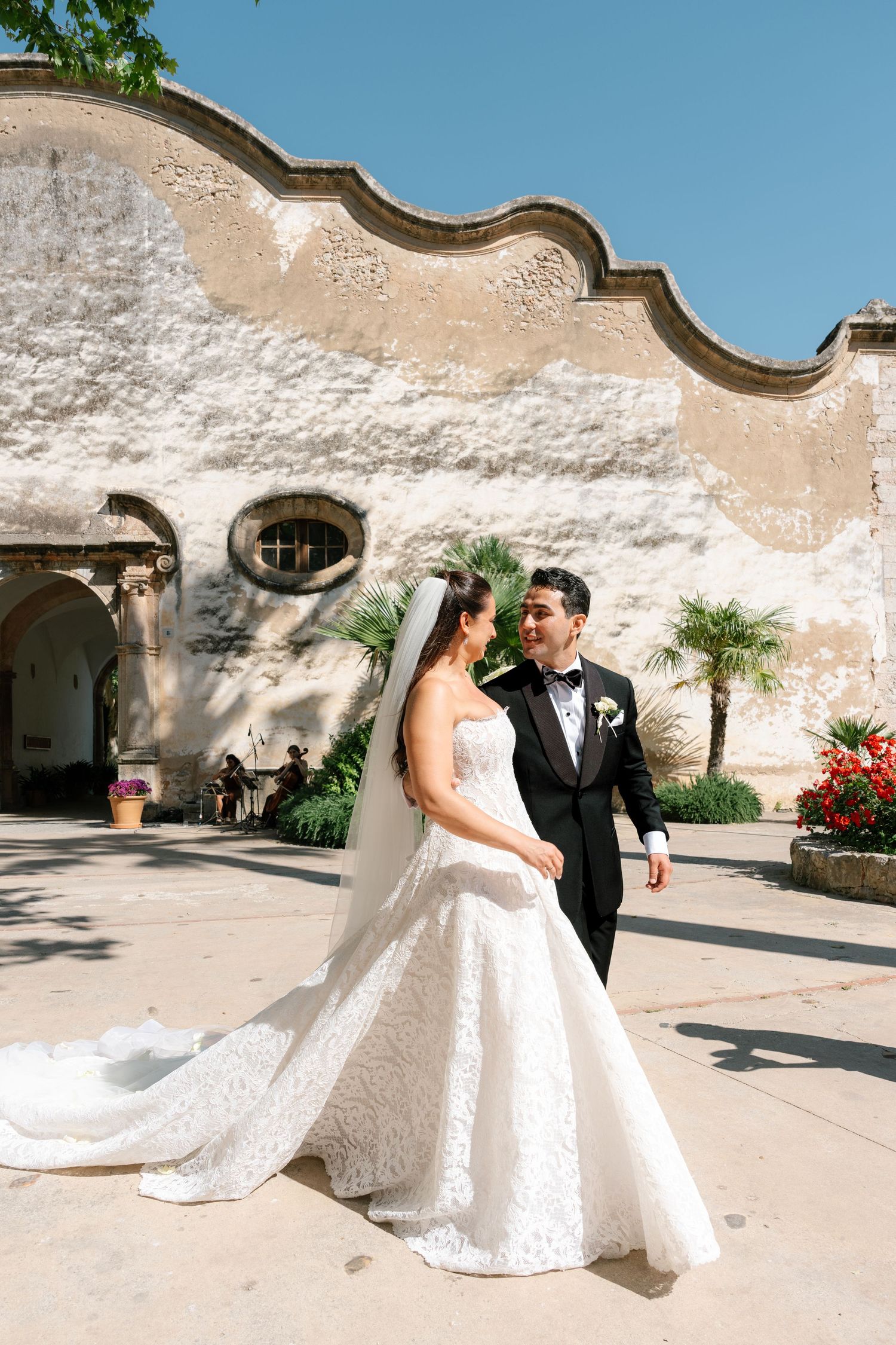 Bride in lace gown and groom in black tuxedo share a romantic moment in a sunlit historic courtyard.