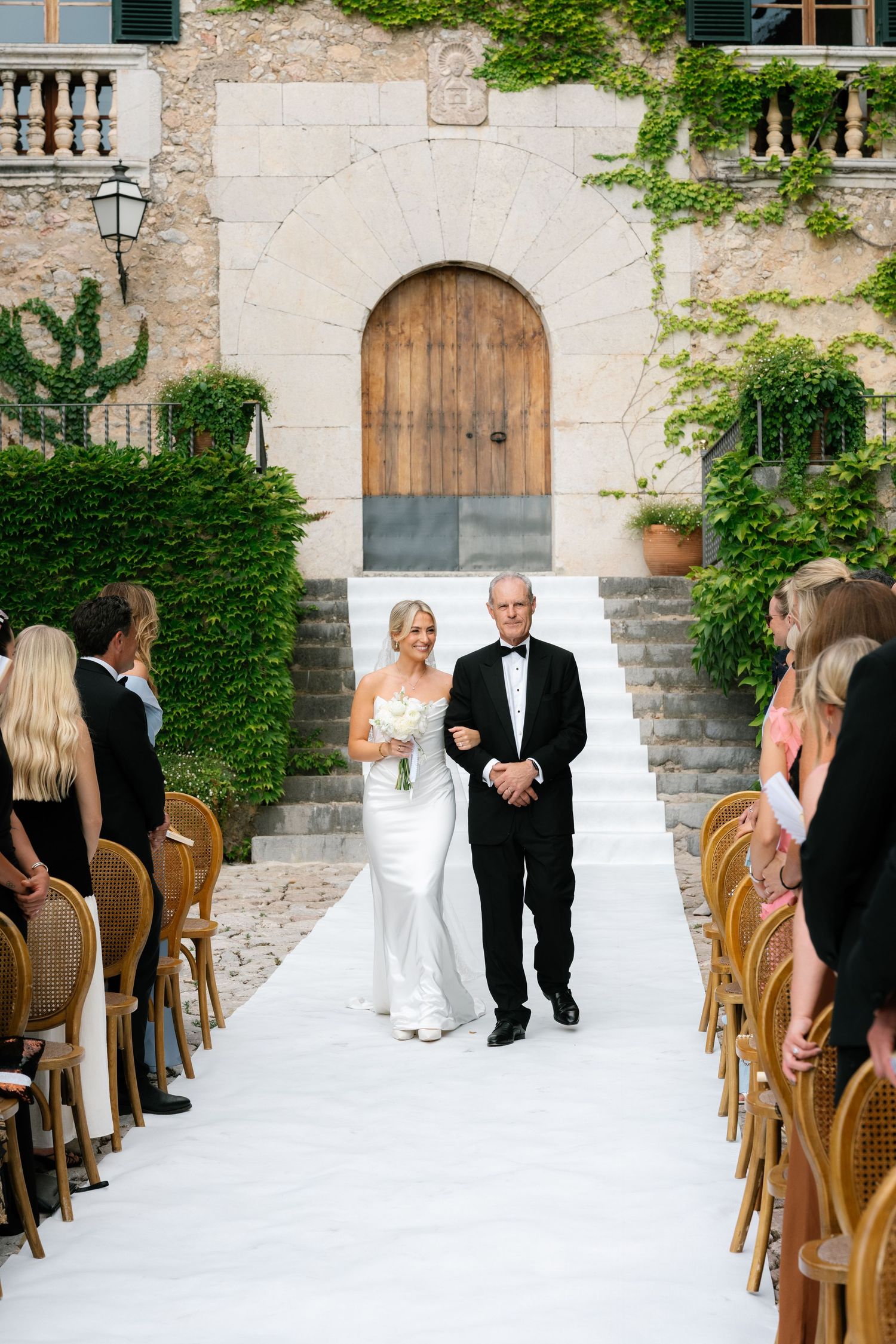 Bride walks down outdoor aisle with escort at elegant stone villa wedding ceremony, flanked by seated guests.