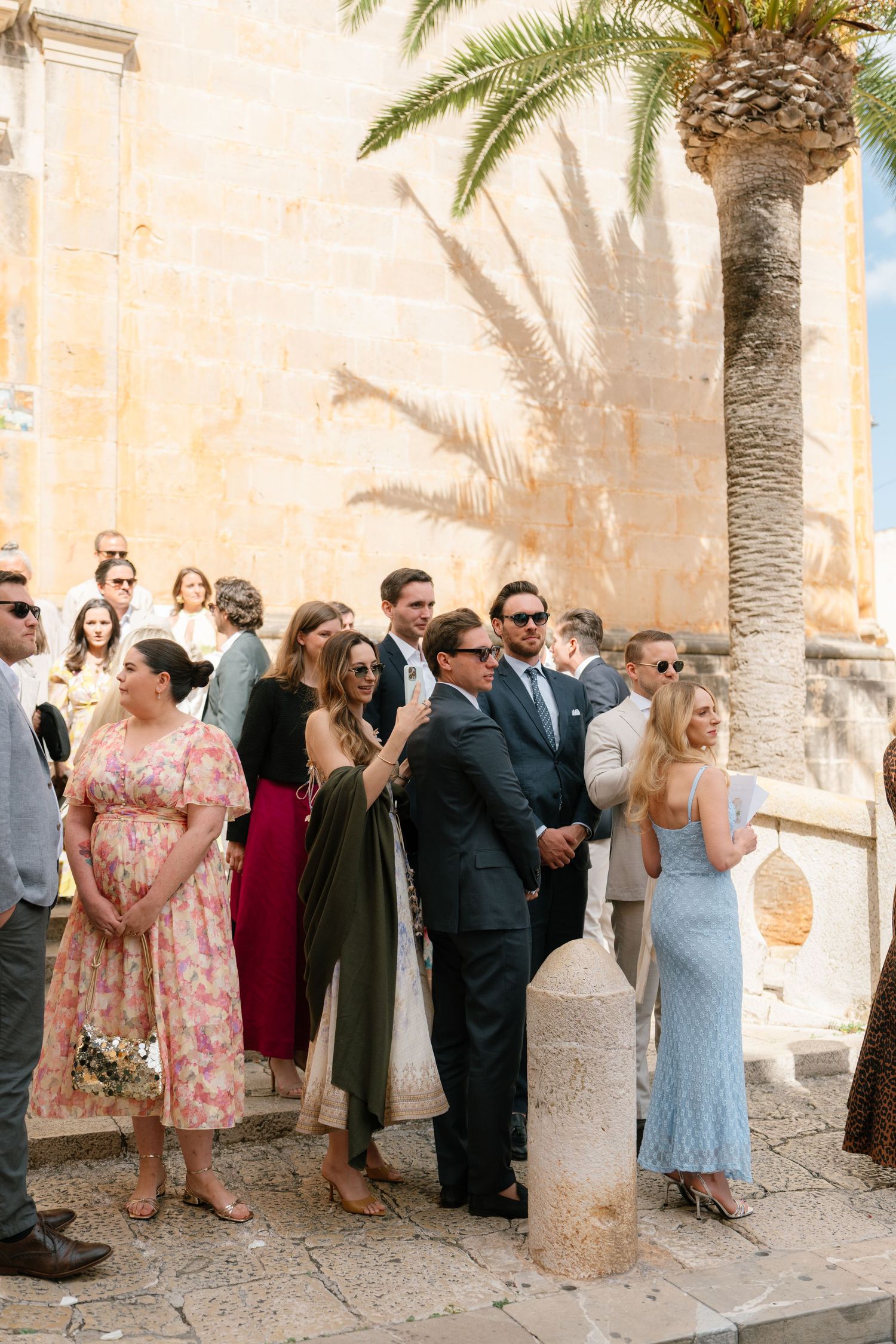 Wedding guests mingle and socialize in an elegant outdoor courtyard with palm trees and stone walls.