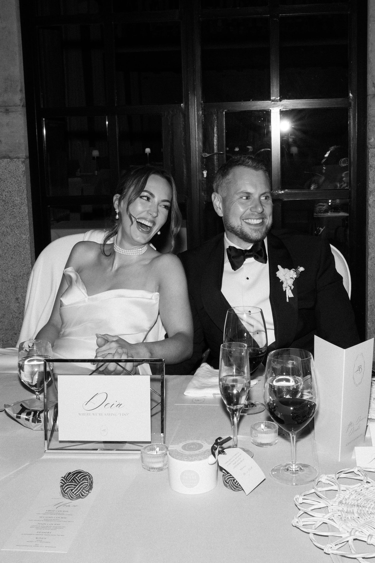 Black and white photo of newlyweds laughing at their reception table with wine glasses and place cards.
