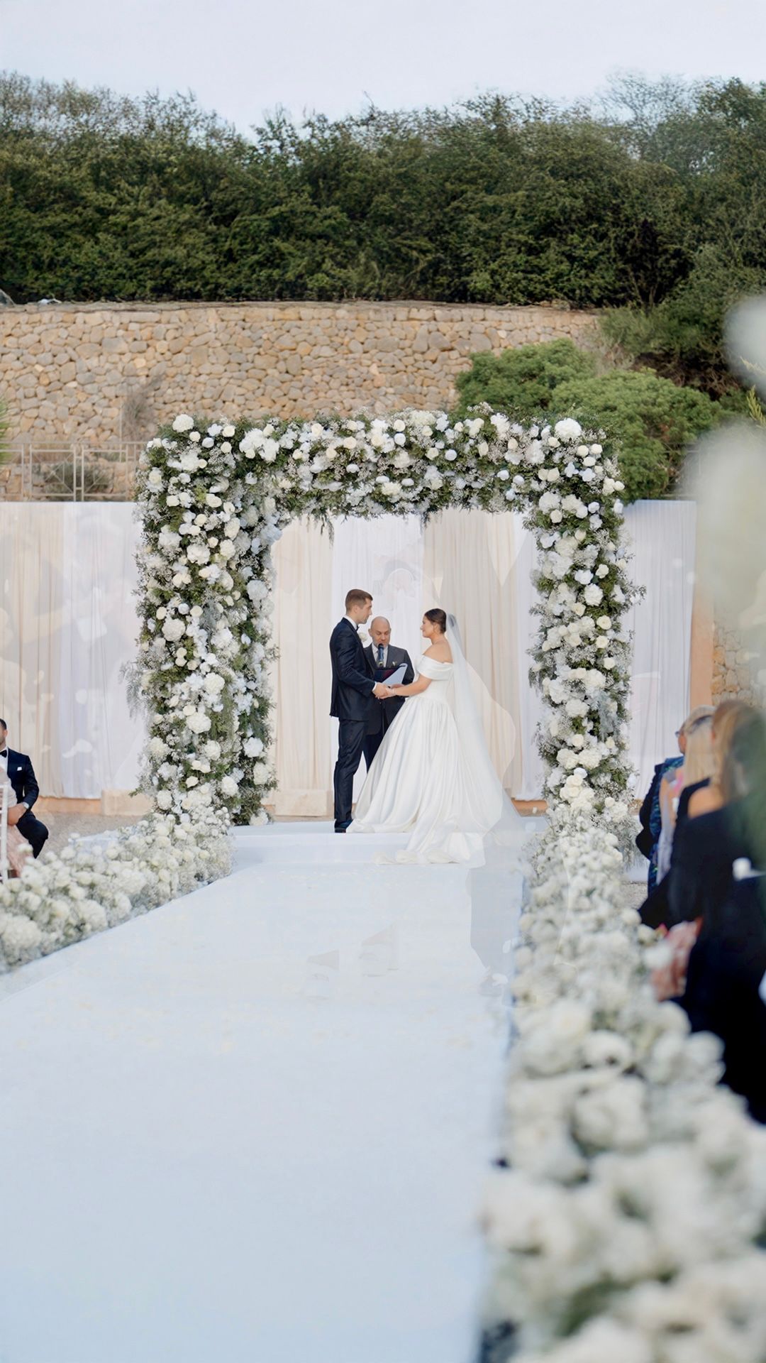 Elegant outdoor wedding ceremony featuring a white floral arch and draped fabric backdrop.