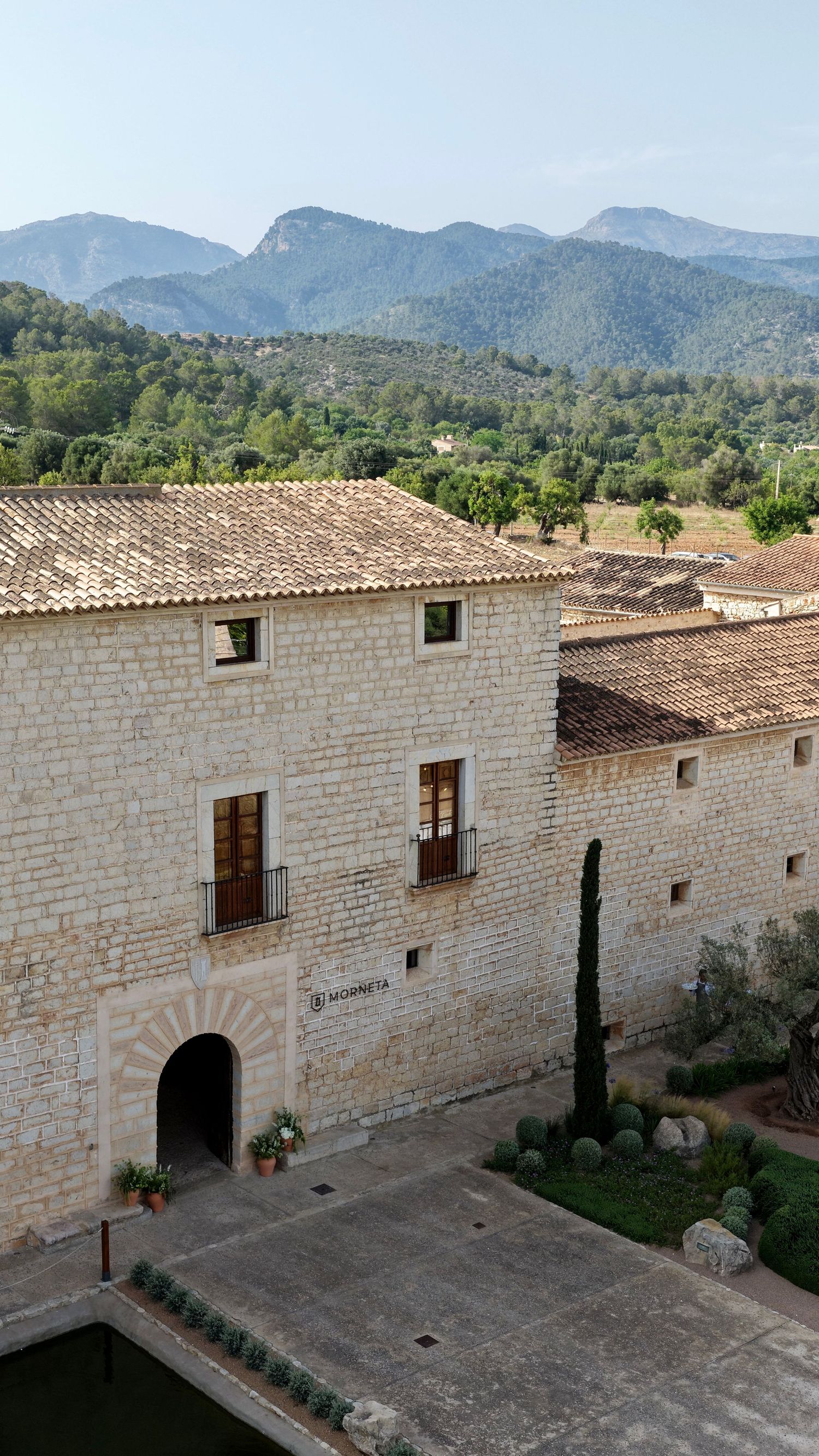 Historic stone building with arched entrance and mountain landscape backdrop in Mediterranean countryside setting.