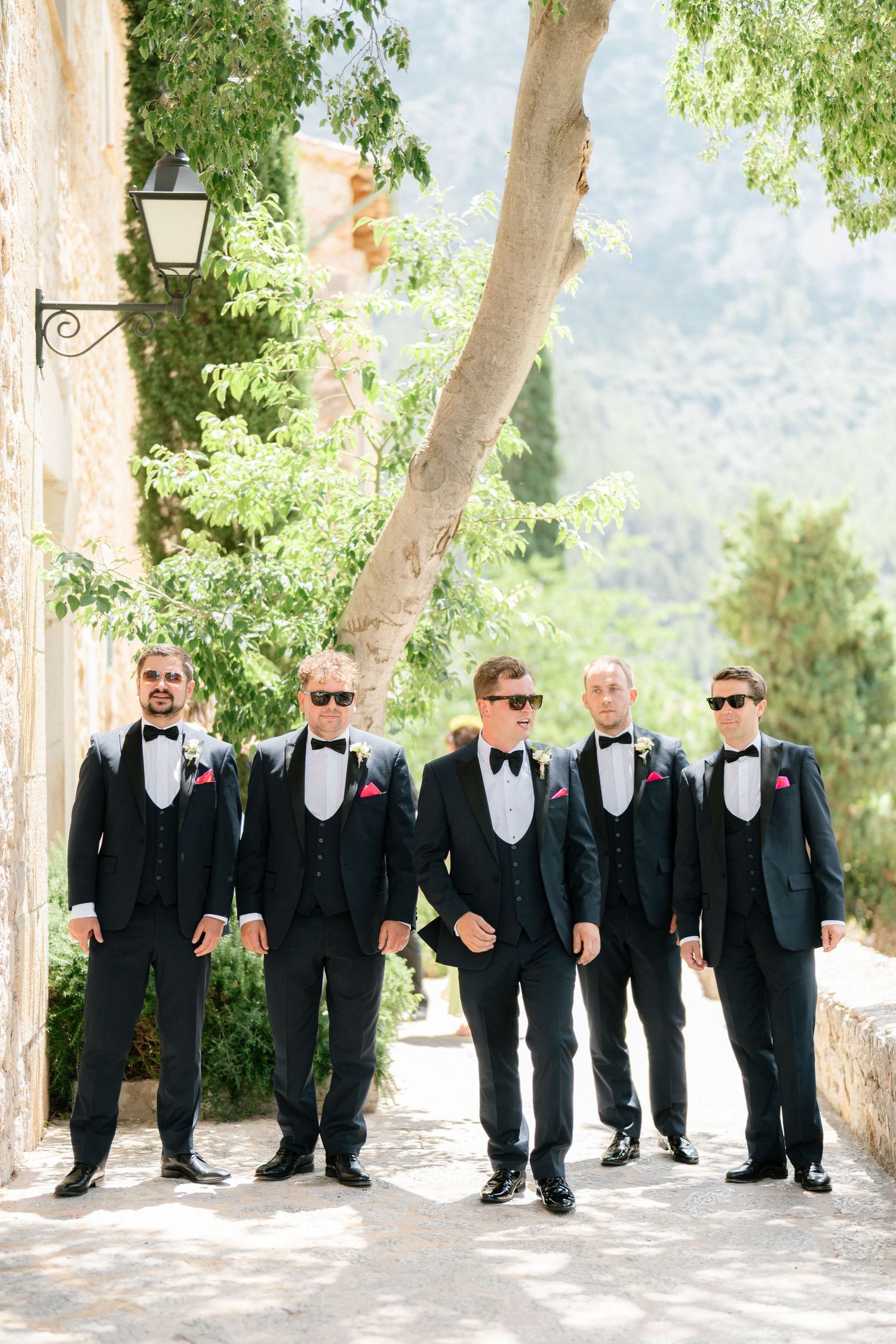 Groomsmen in black tuxedos and sunglasses pose on a stone path with mountain backdrop.