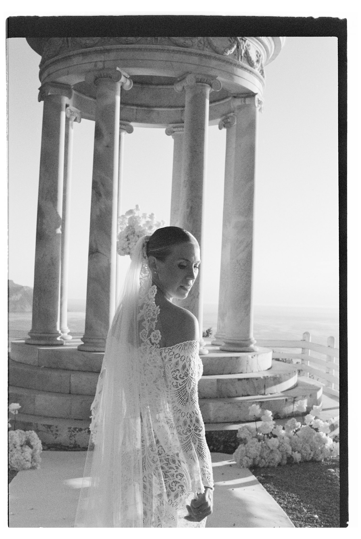 Bride in lace dress posing between elegant stone columns with scenic backdrop.