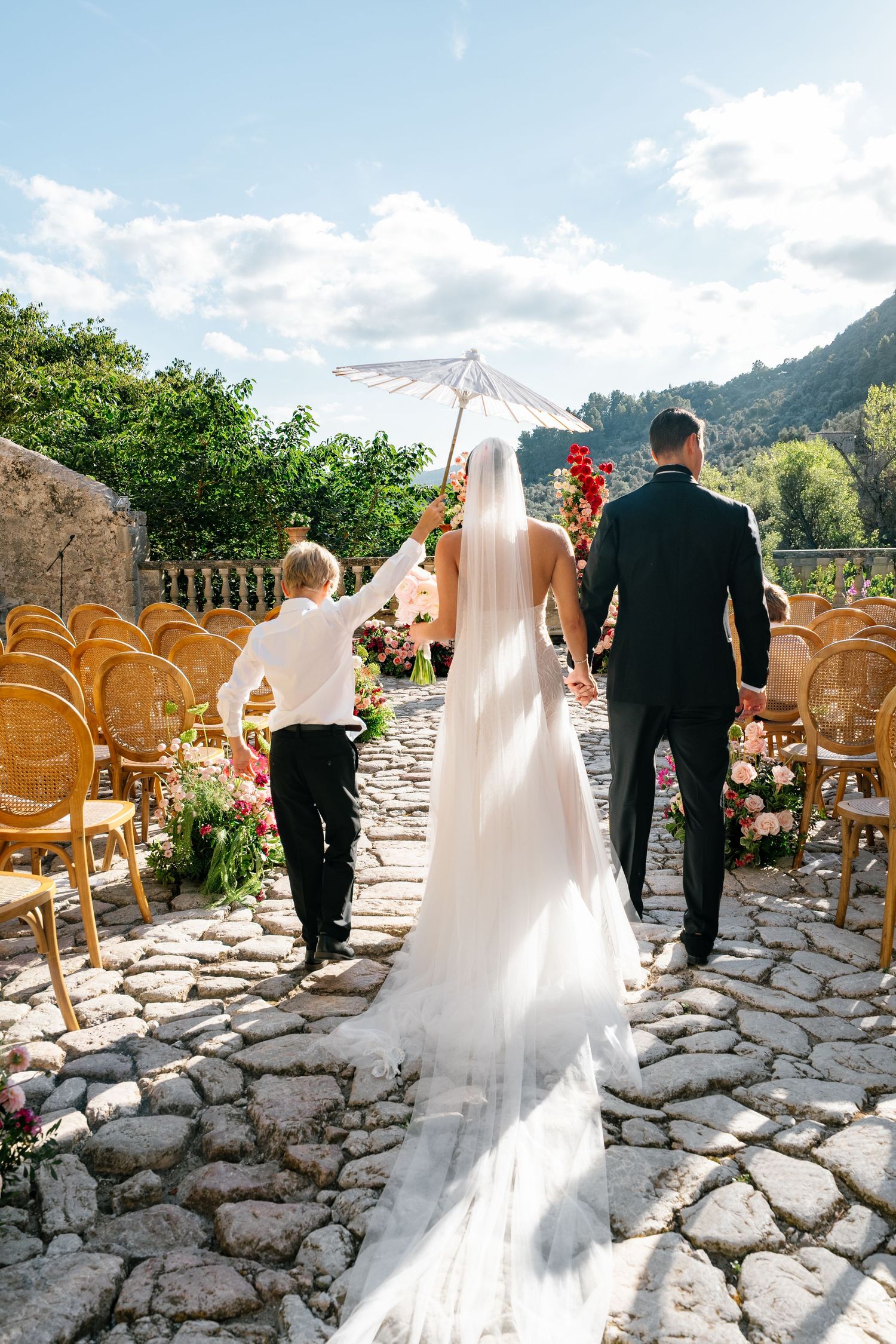 Wedding party walking along stone pathway with white parasols and mountain views.