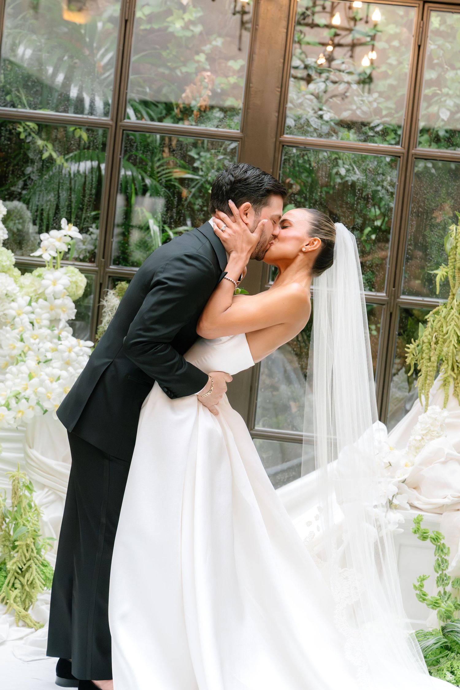 Newlyweds share a romantic kiss in front of a greenhouse window decorated with white orchids and greenery.