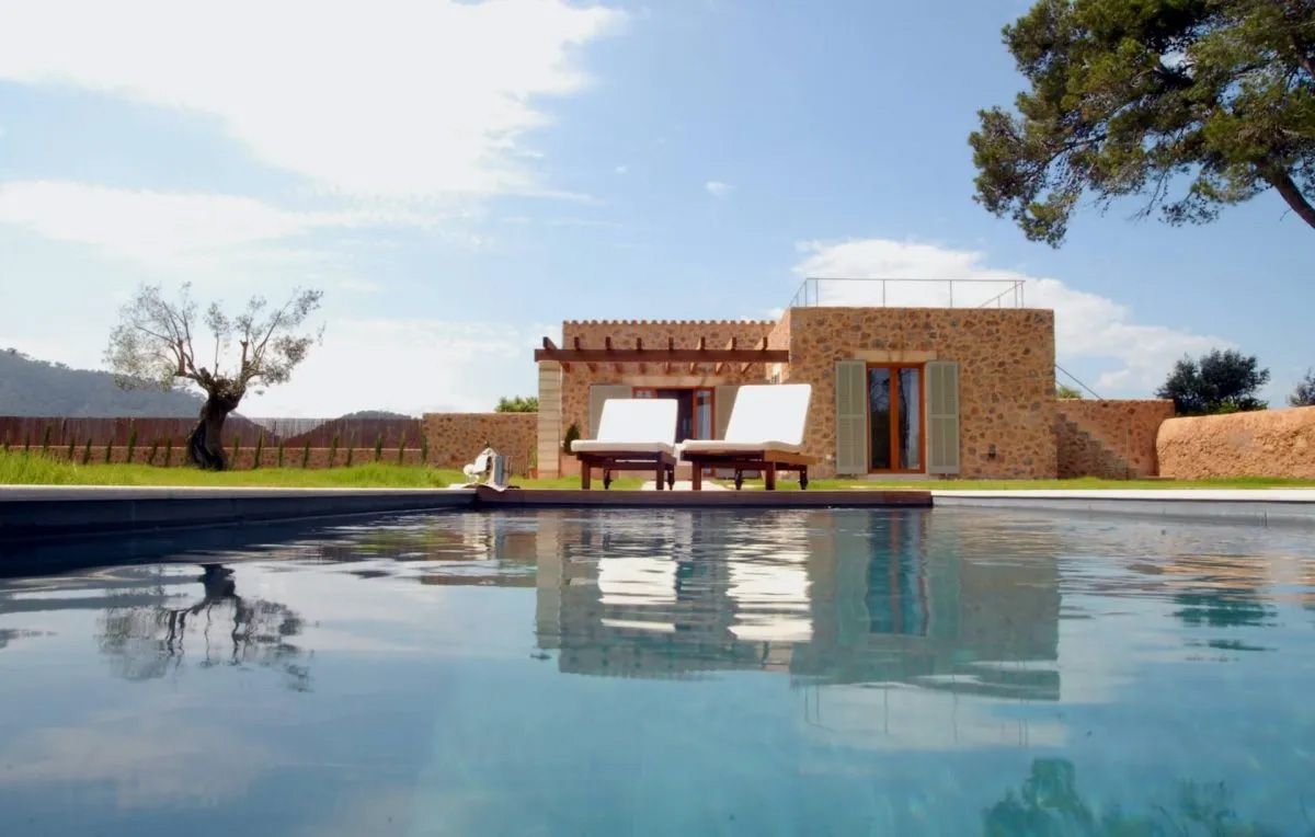 Stone villa Can Simoneta with private pool, sun loungers, and lush trees under a blue sky in a serene Mediterranean setting.