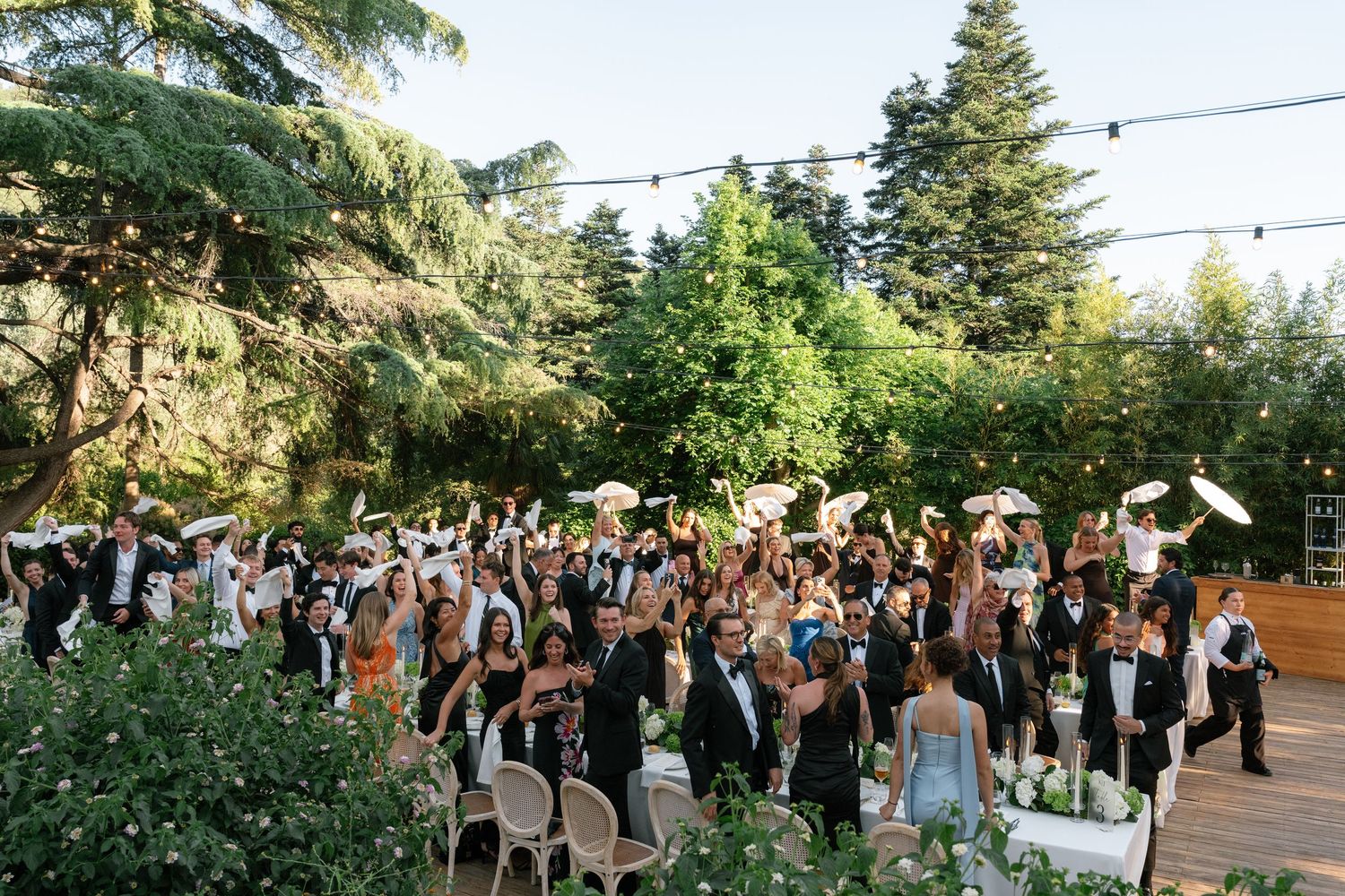 Outdoor wedding reception guests celebrating on a decorated wooden deck surrounded by lush trees and string lights.